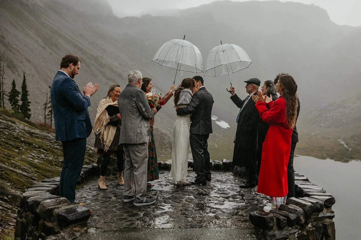 Newly married couple kiss under clear umbrellas as guests applaud during a rainy Washington mountain elopement ceremony with dramatic foggy backdrop.