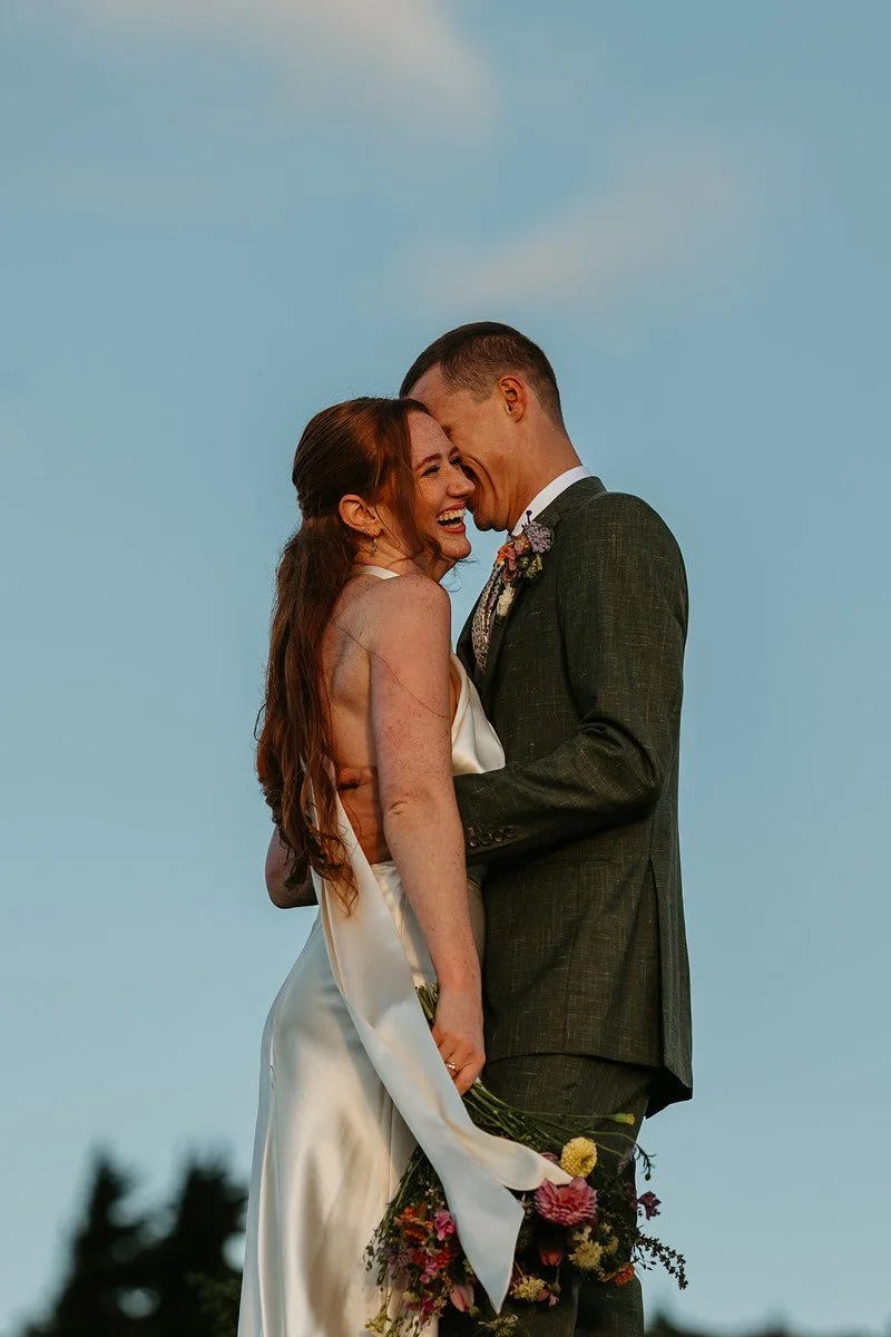 Bride and groom embracing and laughing together at golden hour, bouquet in hand, framed against a clear blue sky.