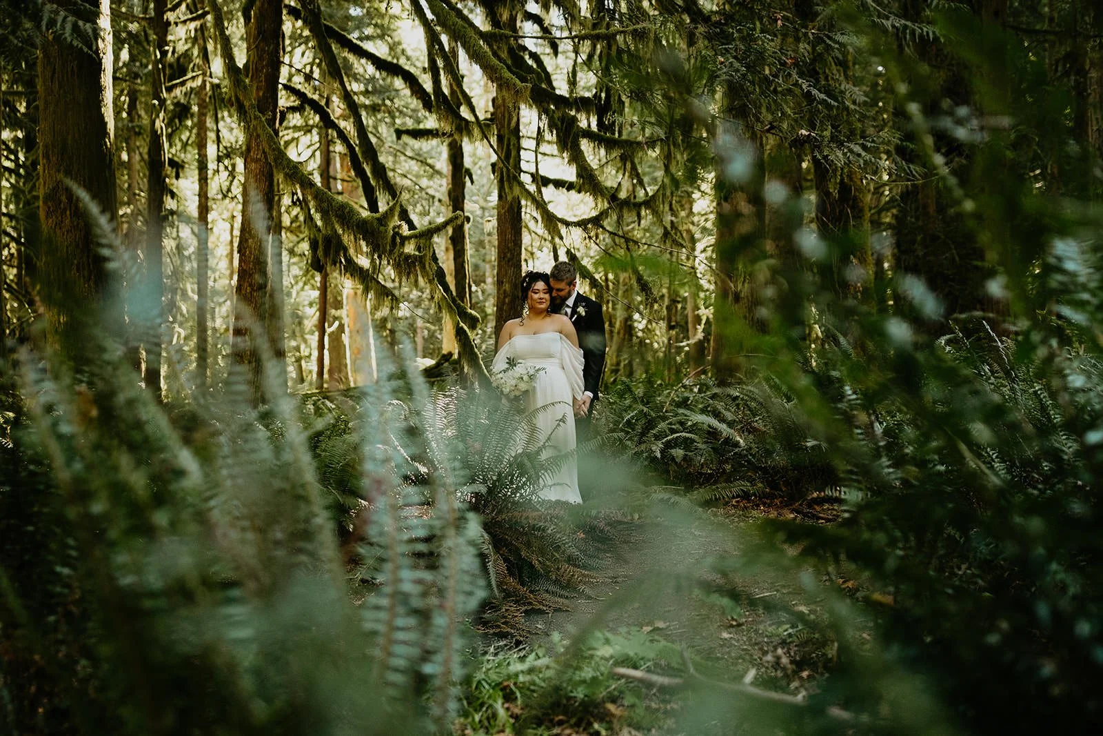 Eloping couple walking hand in hand along a fern-lined forest trail beneath towering trees and hanging moss.