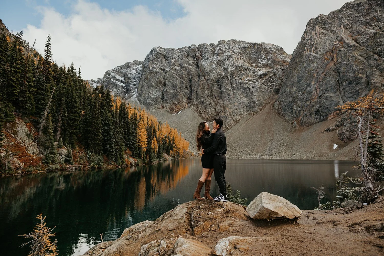 Couple dressed up stand in front of a blue lake with golden larches and jagged mountains in the background