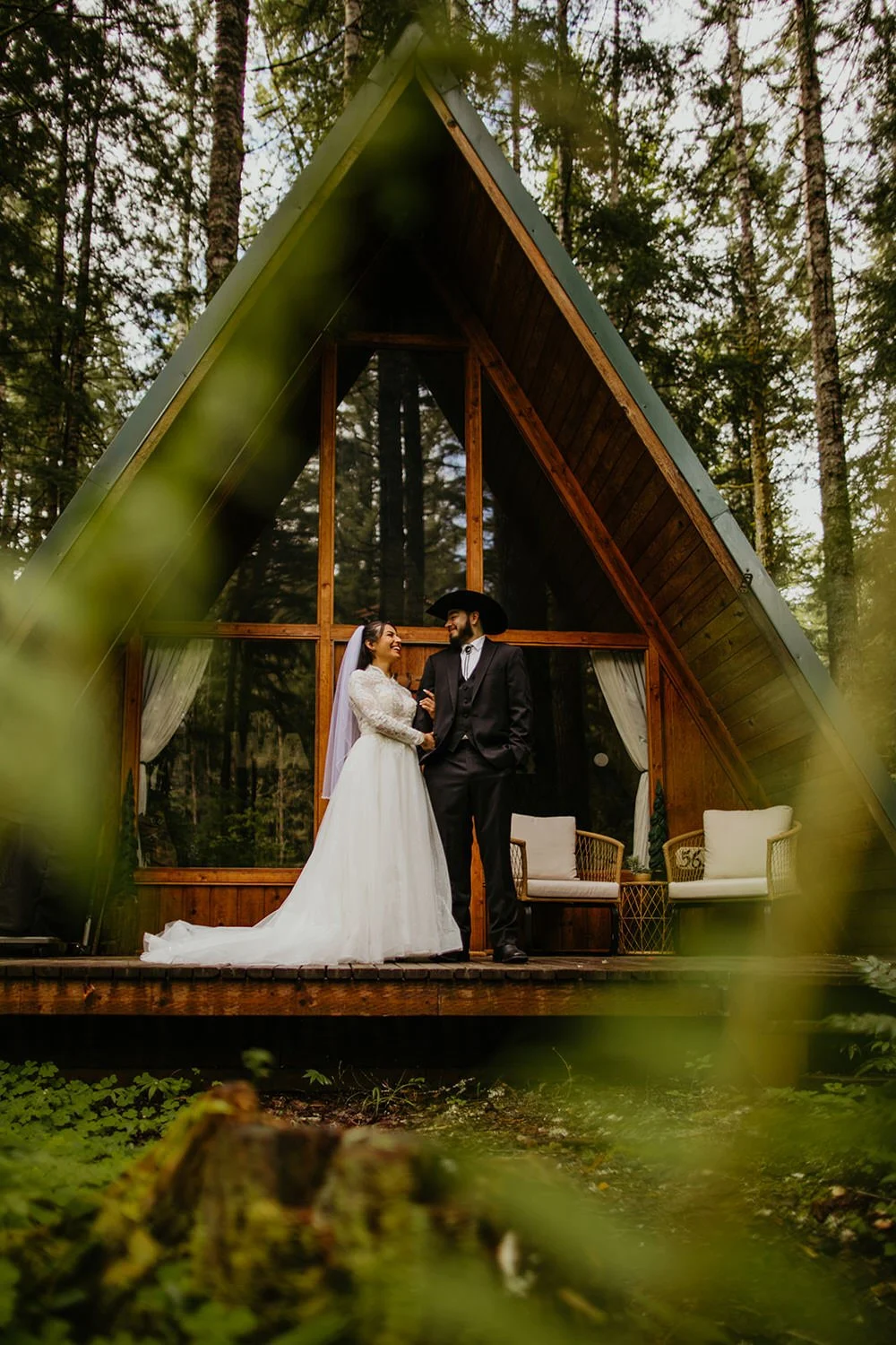 Couple embracing outside a forest A-frame cabin in Washington, photographed through green foliage during their elopement