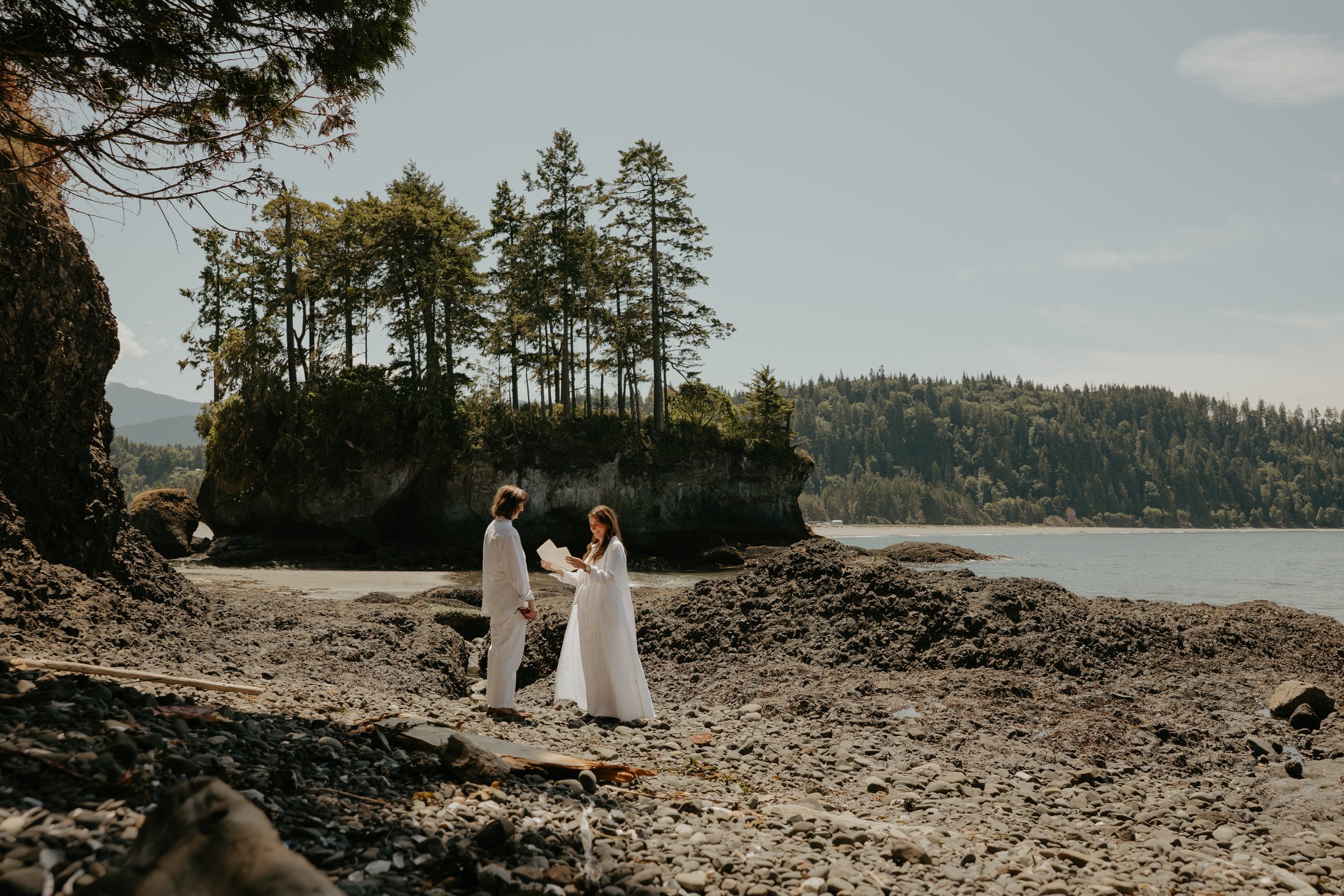 Intimate queer elopement ceremony on a rocky Washington beach with a forested sea stack island and calm water in the background.