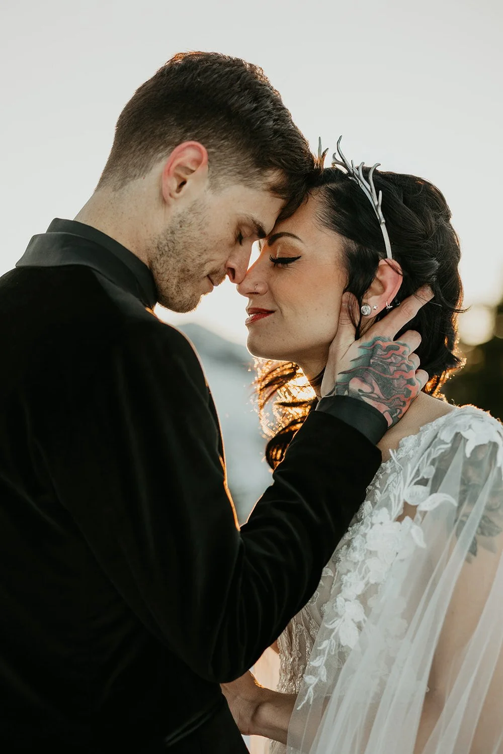 A newlywed couple with foreheads touching, eyes closed, during sunset. The groom is holding the bride's face, and they are dressed in wedding attire. The bride has dark hair, is wearing a tiara, earrings, and a lace wedding dress, while the groom has tattoos visible on his hand and is wearing a black suit.
