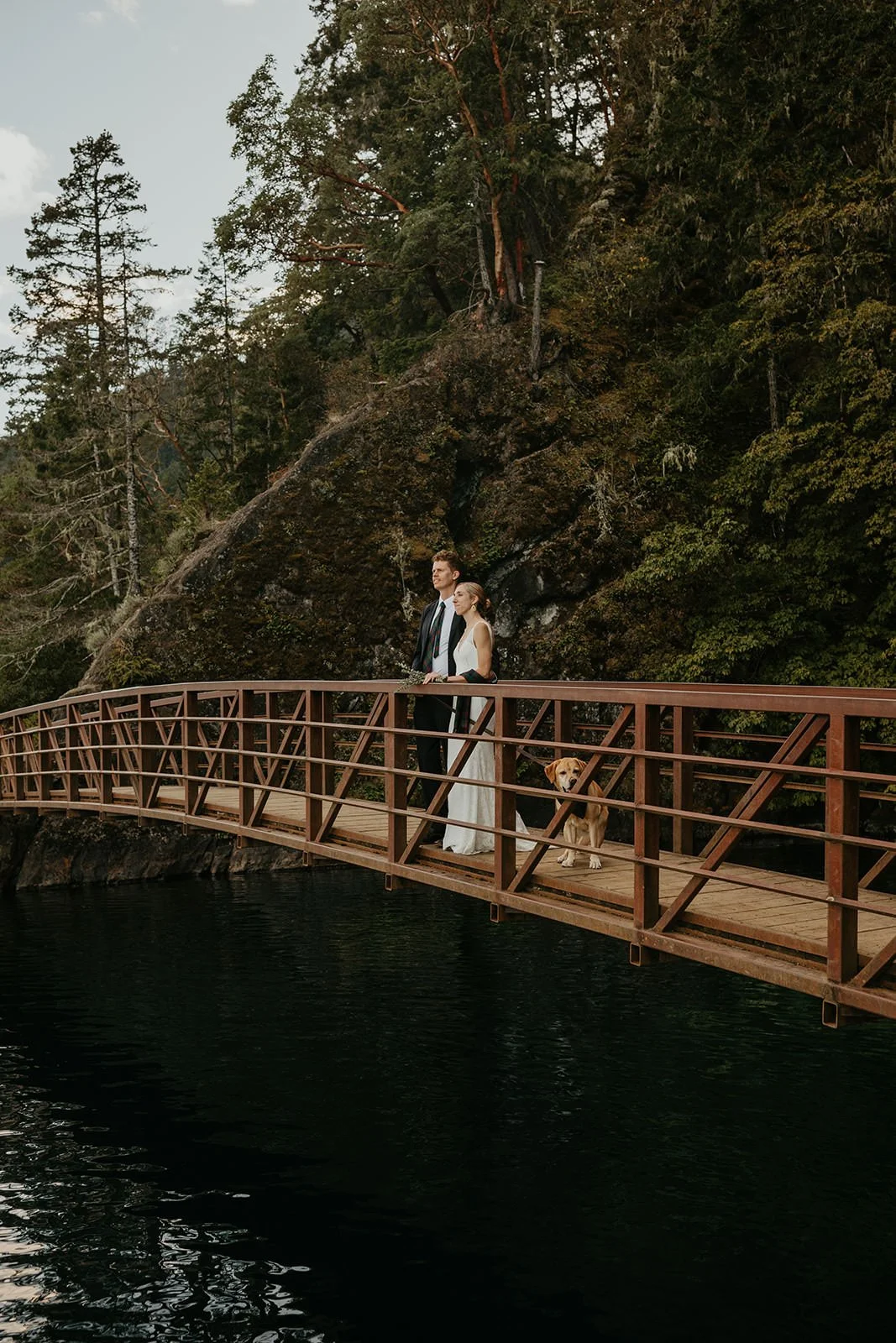Couple walking across a wooden footbridge above a dark, still river, surrounded by steep forested cliffs.