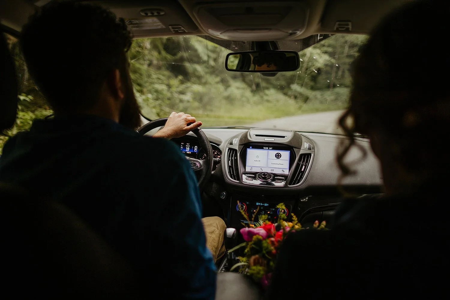 View from inside a car driving a winding forest road, with flowers resting near the center console.