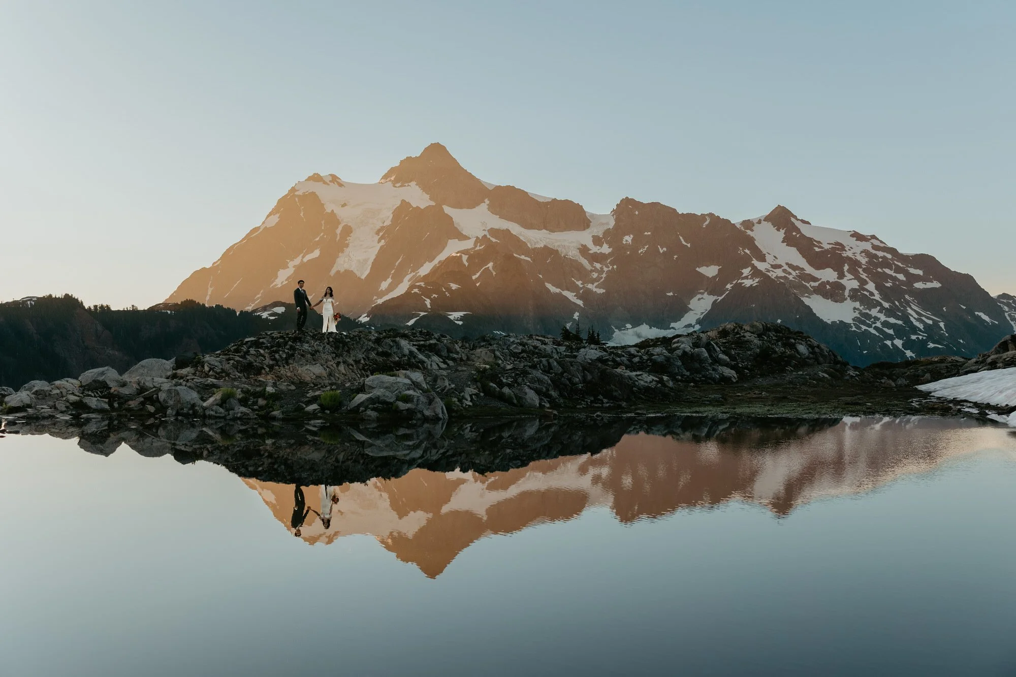 Elopement couple reflected in an alpine lake at sunrise, with rugged mountain peaks mirrored in calm water.