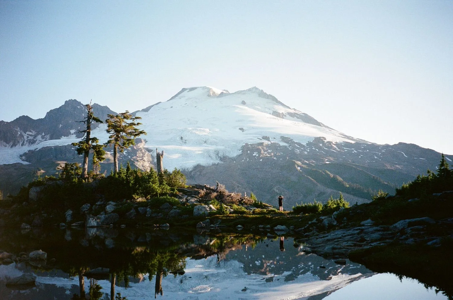 A mountain with snow and glaciers, green trees, rocks, and water reflecting the landscape, with a person standing near the water.