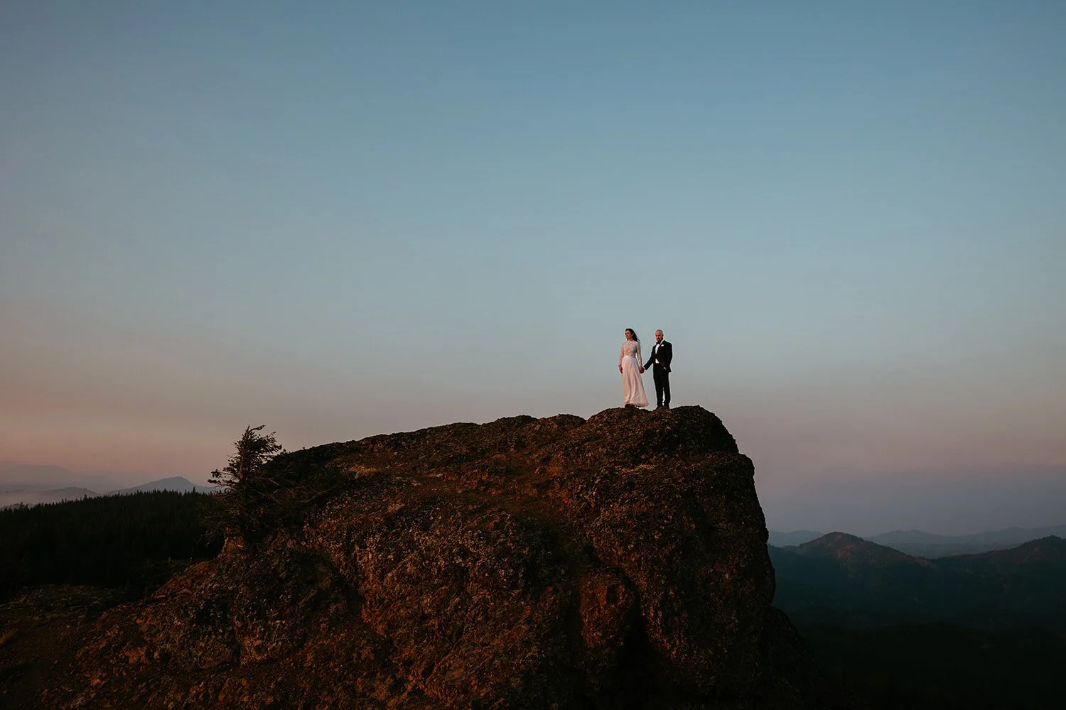Couple standing on the edge of a rocky cliff at dusk with soft pastel sky and layered mountain silhouettes in the distance
