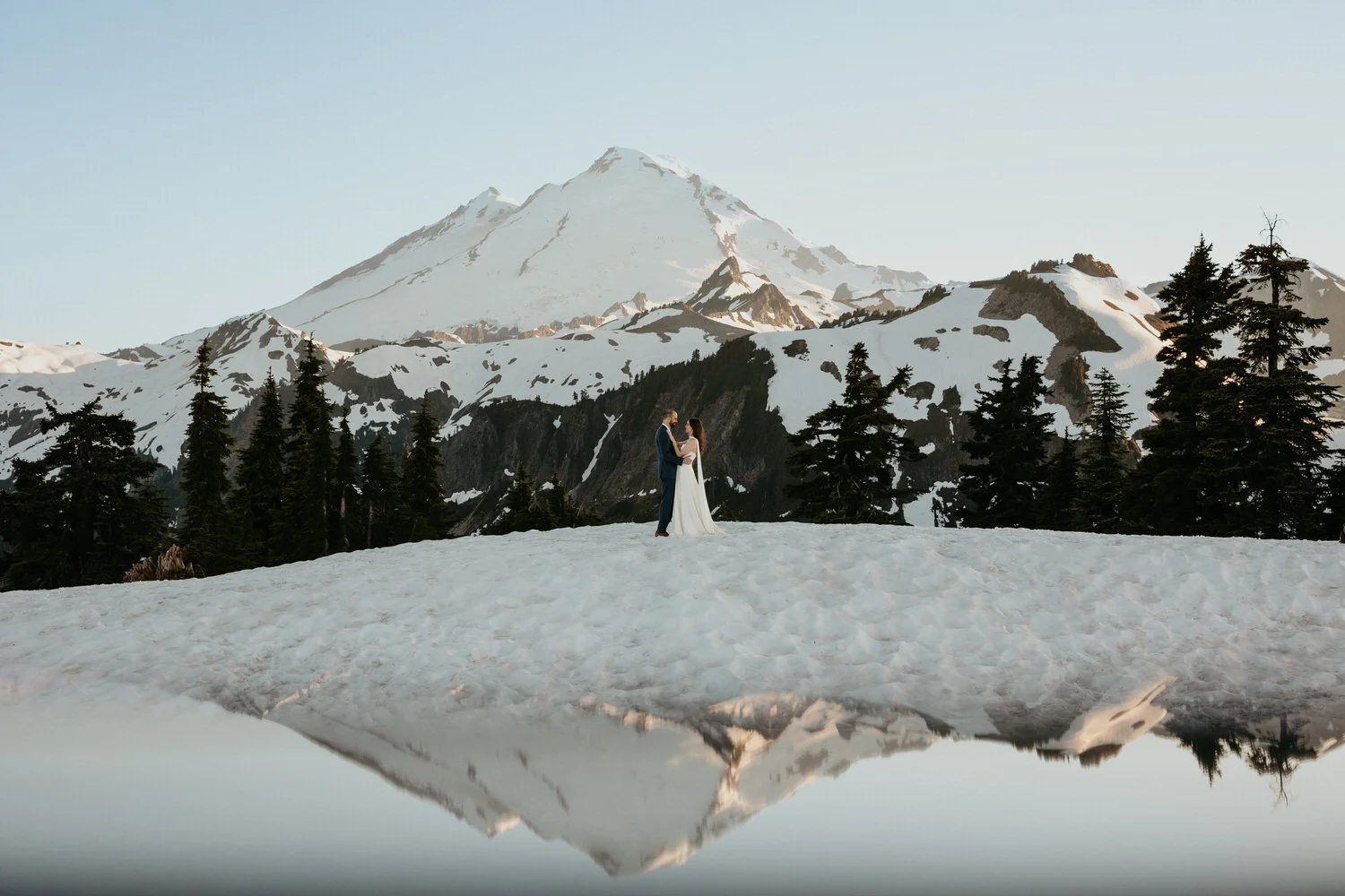 Couple standing together on a snowy alpine ridge with Mount Baker reflected in a still mountain lake at sunset.
