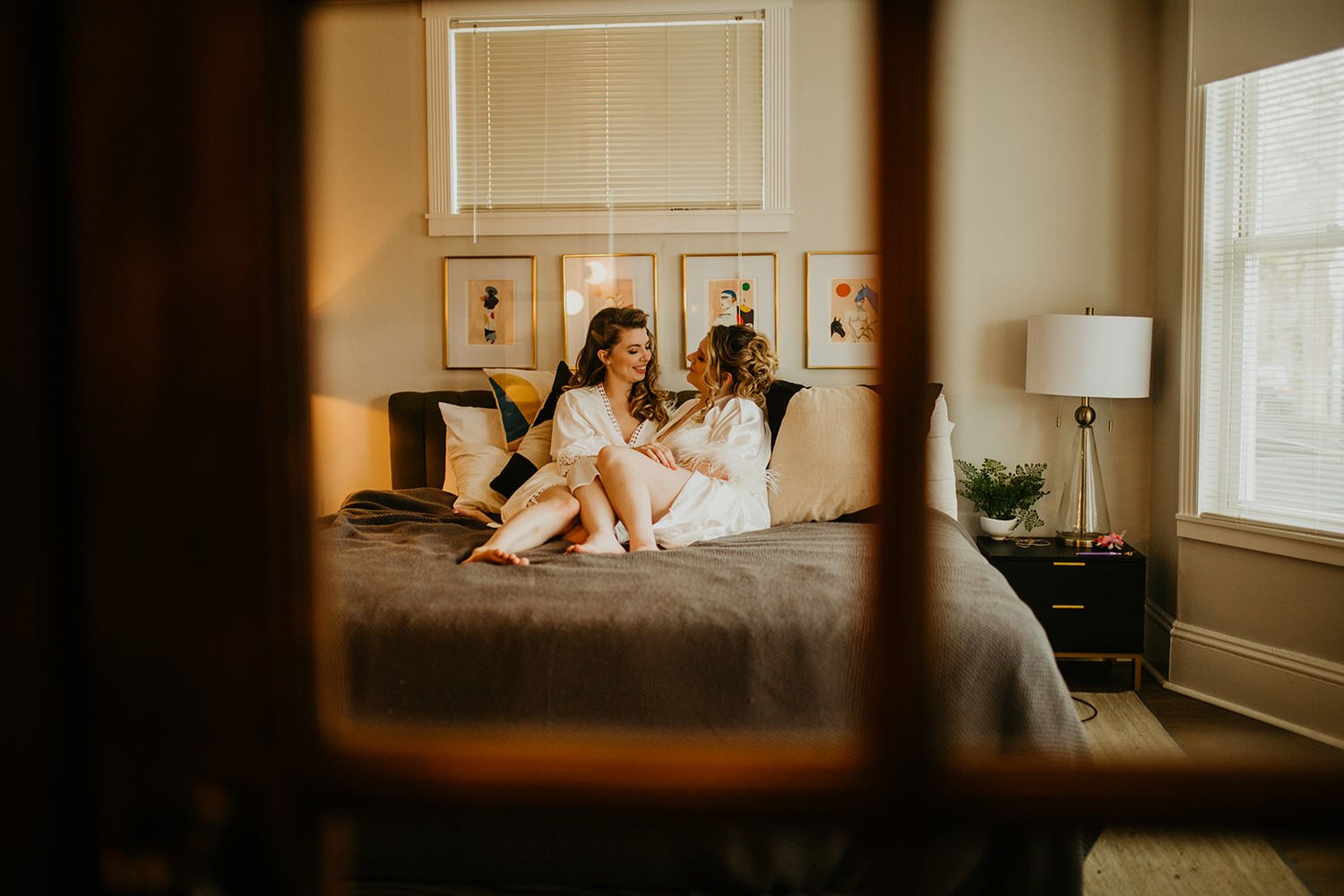 Two brides sitting together on a bed in a cozy hotel room, wearing matching white robes and sharing a quiet, intimate moment before their wedding.