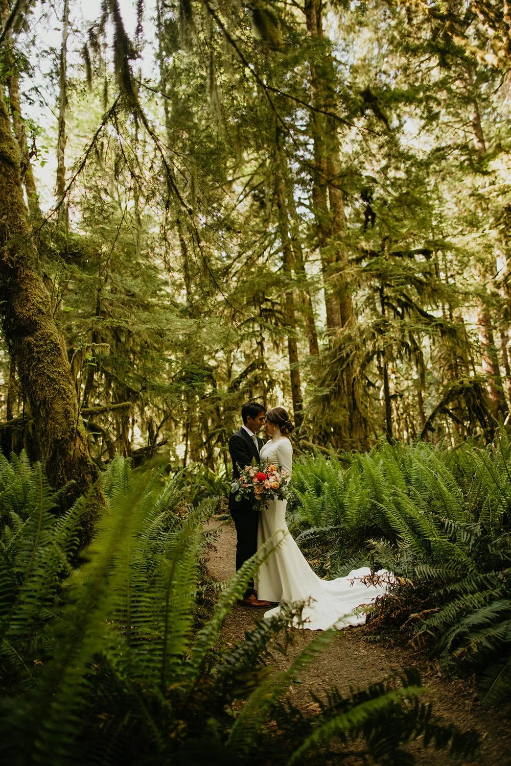 Couple standing together on a forest trail surrounded by lush ferns during a moody Washington elopement