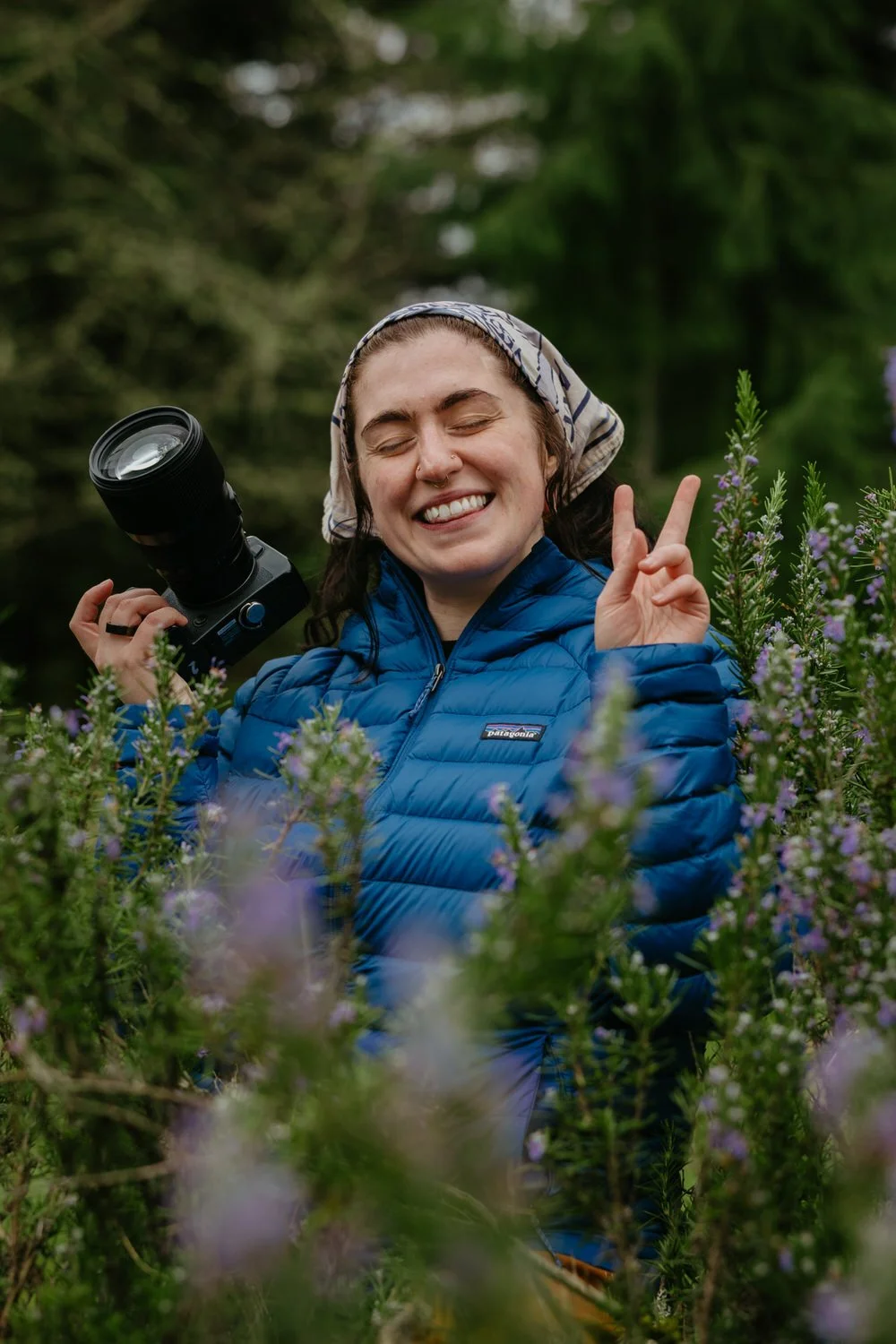 A woman with a camera, wearing a blue puffer jacket and a headscarf, smiling and making a peace sign while standing in a field of purple flowers.