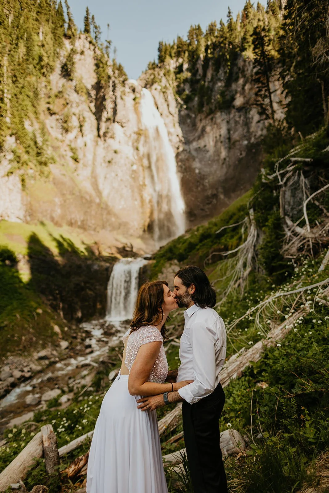 Couple kissing in front of a tall waterfall in Washington during their elopement, surrounded by cliffs, moss, and flowing water