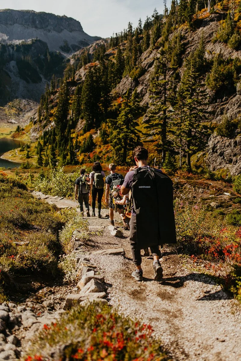 Couple and guests hiking an alpine trail in Washington while carrying a suit garment bag and florals for their elopement ceremony.