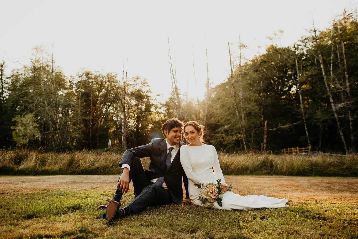 Couple sitting together in an open grassy field at sunset, trees lining the edge of the meadow as warm light falls across the scene.