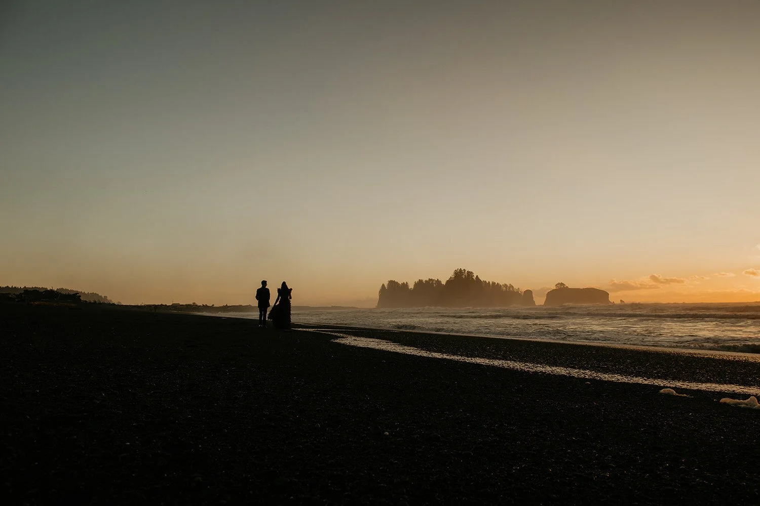 Silhouetted eloping couple standing on a dark beach at sunset, facing the ocean as warm light glows across the horizon.