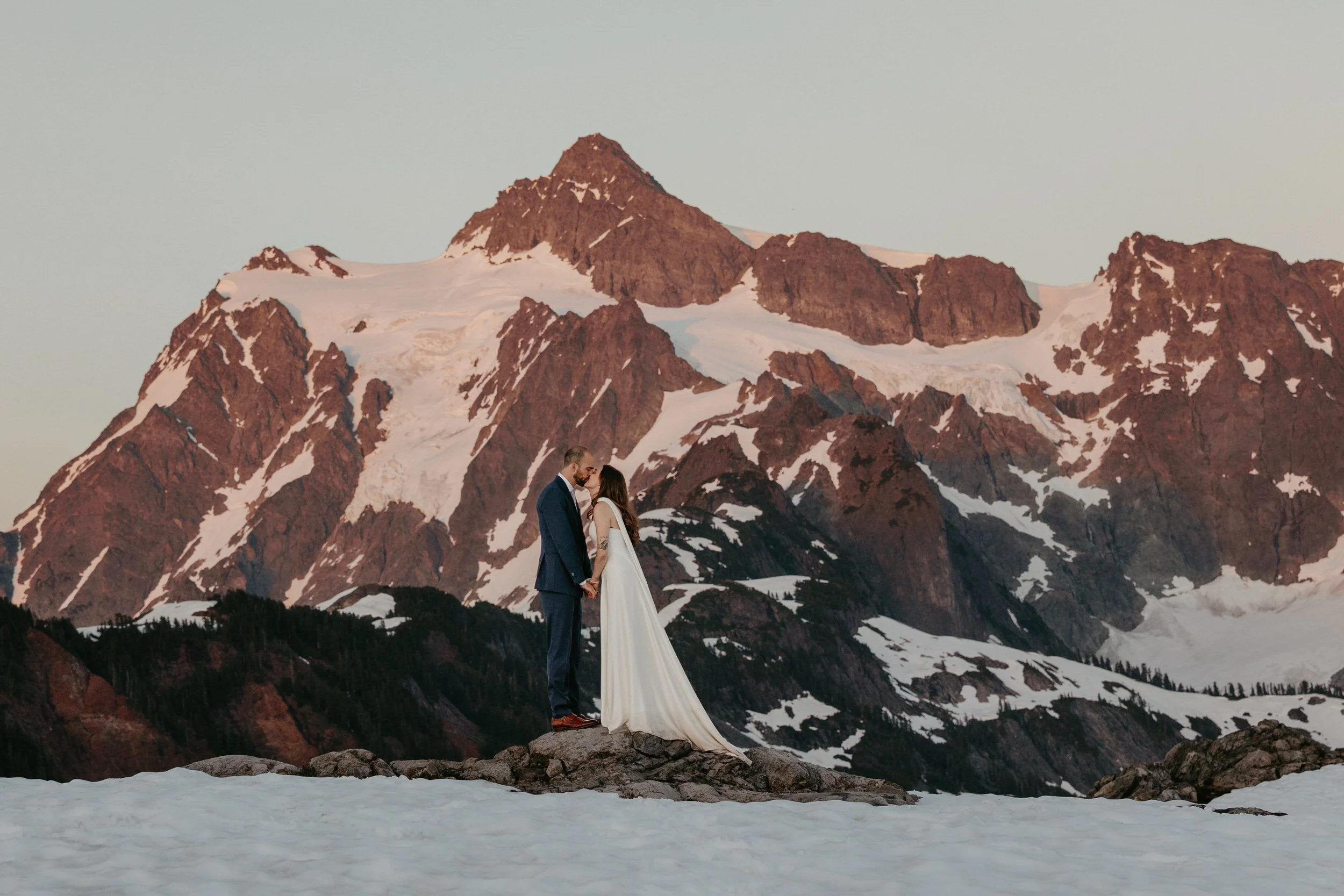 Couple standing on a rocky outcrop as soft pink alpenglow washes over snow-covered peaks in Washington during a mountain elopement.