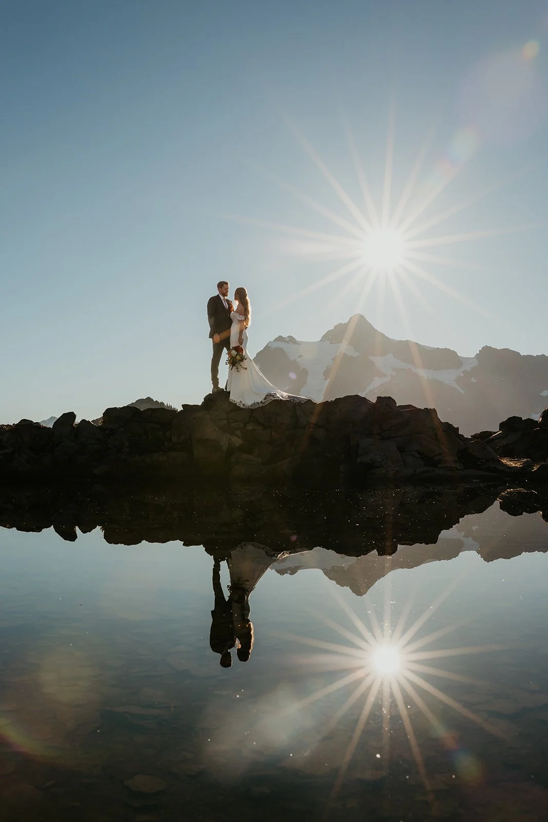A couple stands on a rocky alpine shoreline at sunrise, reflected in a calm mountain lake with jagged peaks silhouetted against the sky.