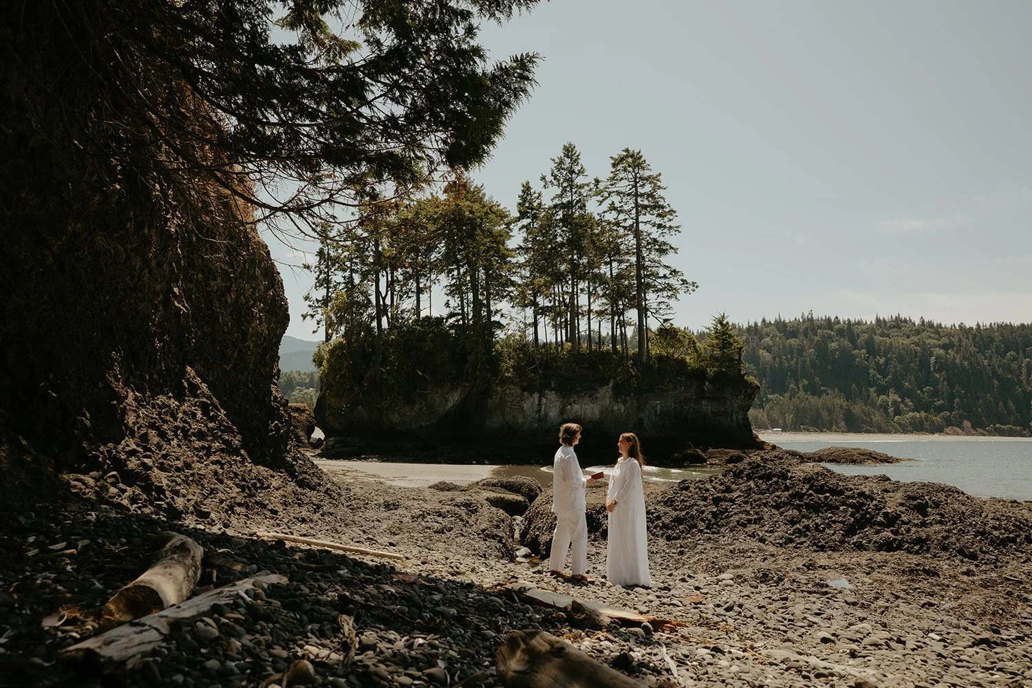 A couple dressed in white, standing on a rocky beach, exchanging vows with a backdrop of trees on an island and ocean under a clear sky.