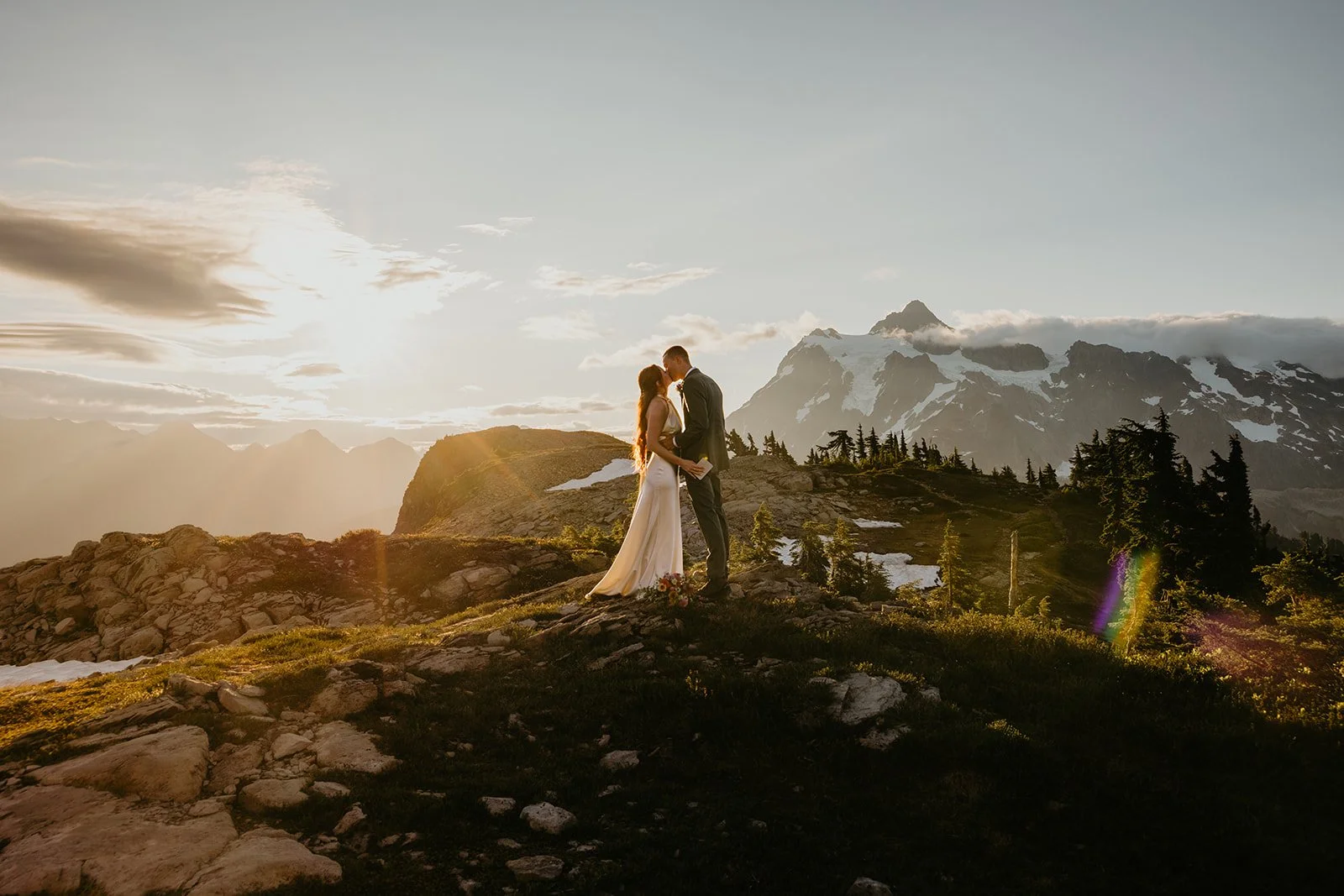 Couple standing together on a grassy bluff during sunset with warm light illuminating Mount Shuksan behind them.
