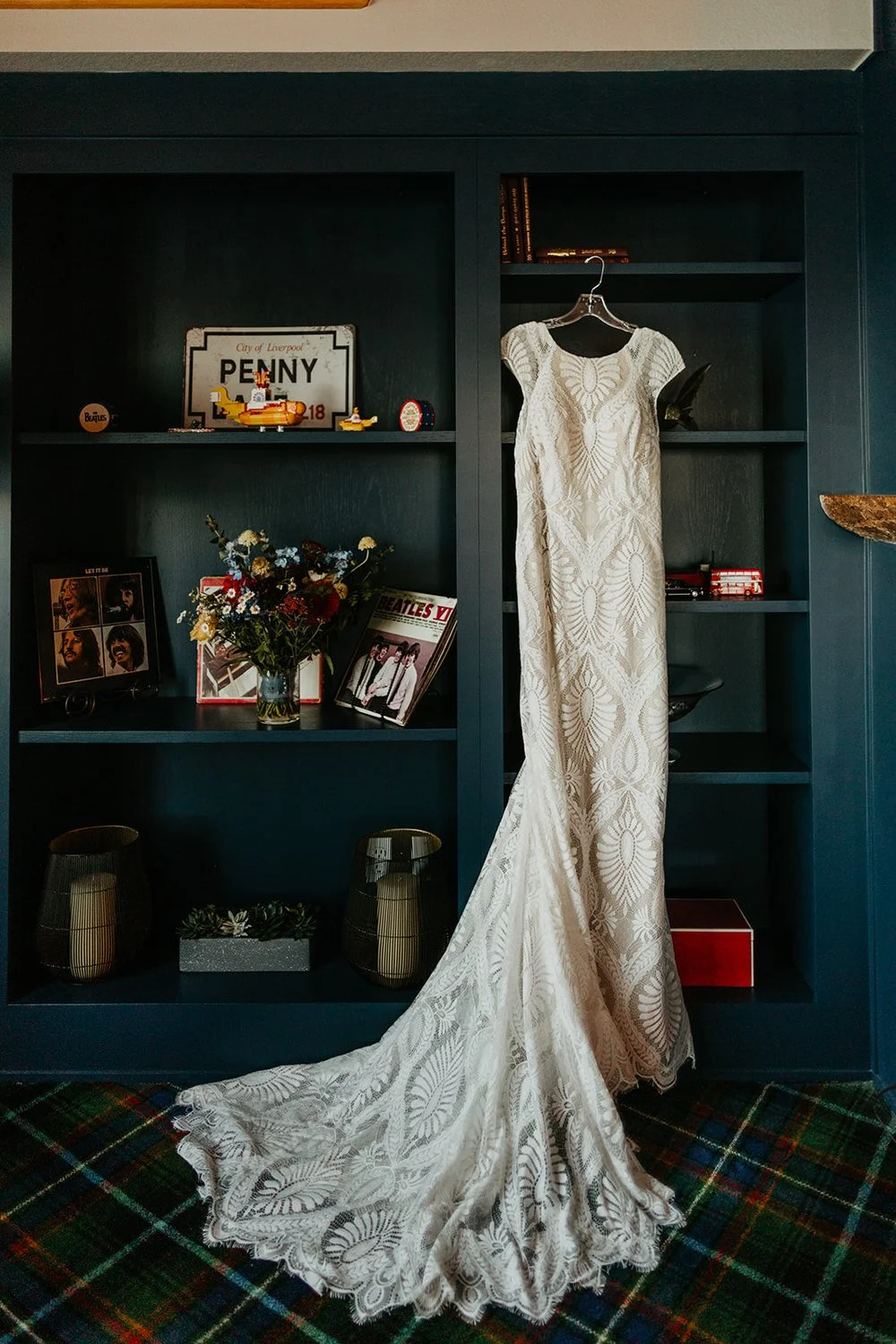 Lace wedding dress hanging on a blue bookshelf decorated with Beatles memorabilia, wildflower bouquet, and personal keepsakes inside a Washington cabin rental.