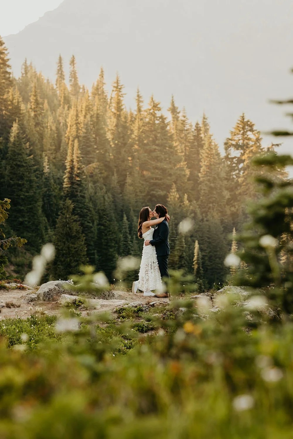 Couple embracing in a quiet alpine meadow surrounded by wildflowers and glowing evergreen forest