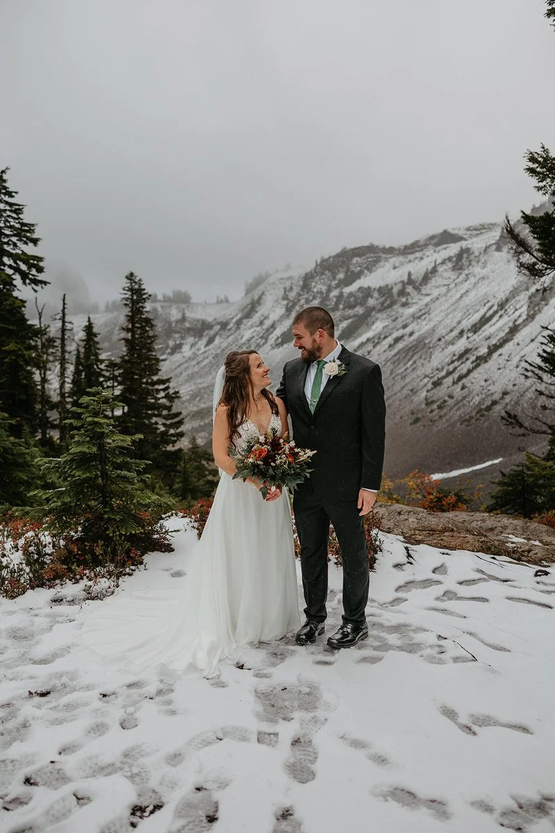 Bride and groom standing together in fresh snow on a mountain overlook, smiling at each other with evergreen trees and foggy peaks in the background.
