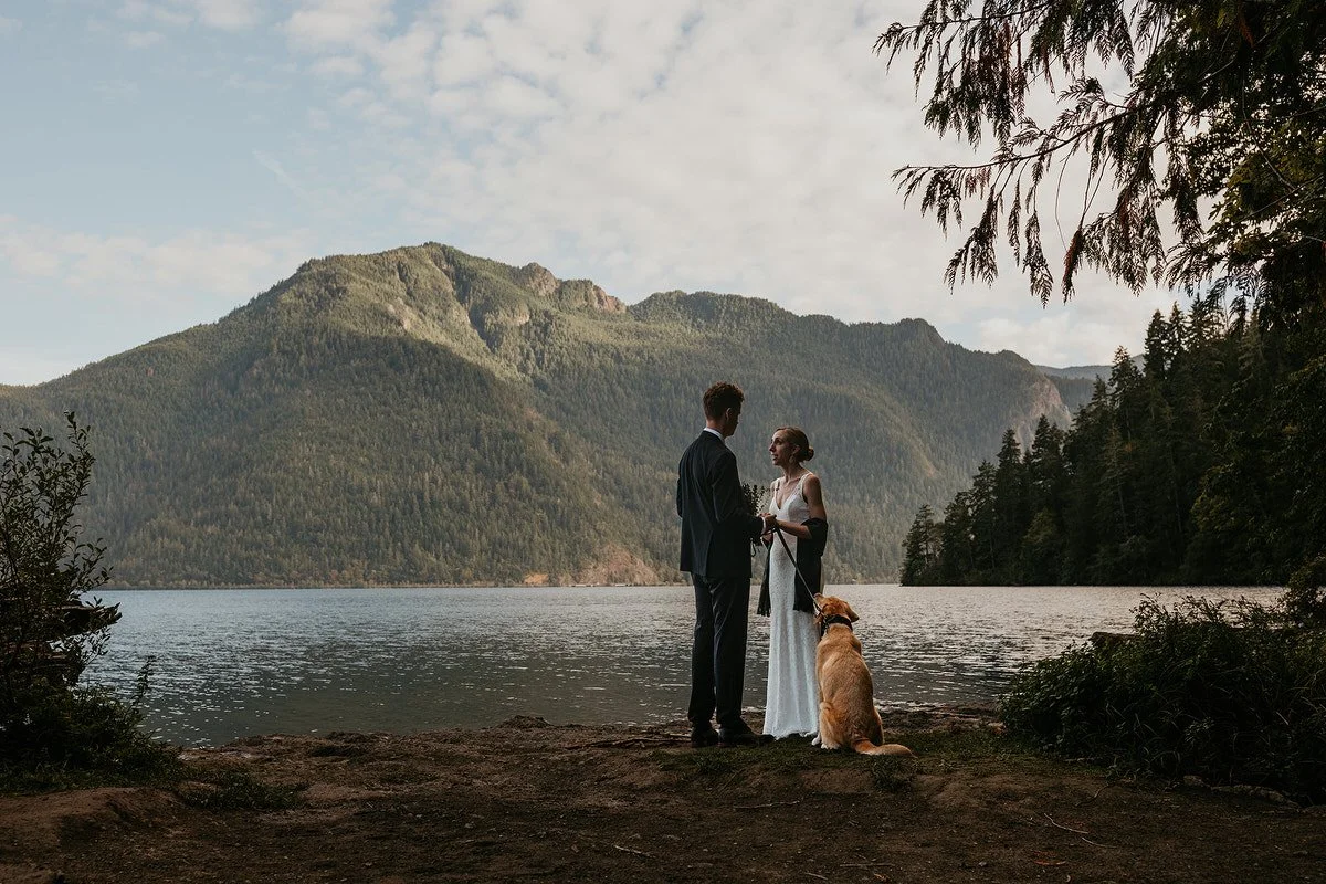 Couple exchange vows beside a peaceful Washington lake with their dog sitting between them, framed by evergreen trees and mountain views.