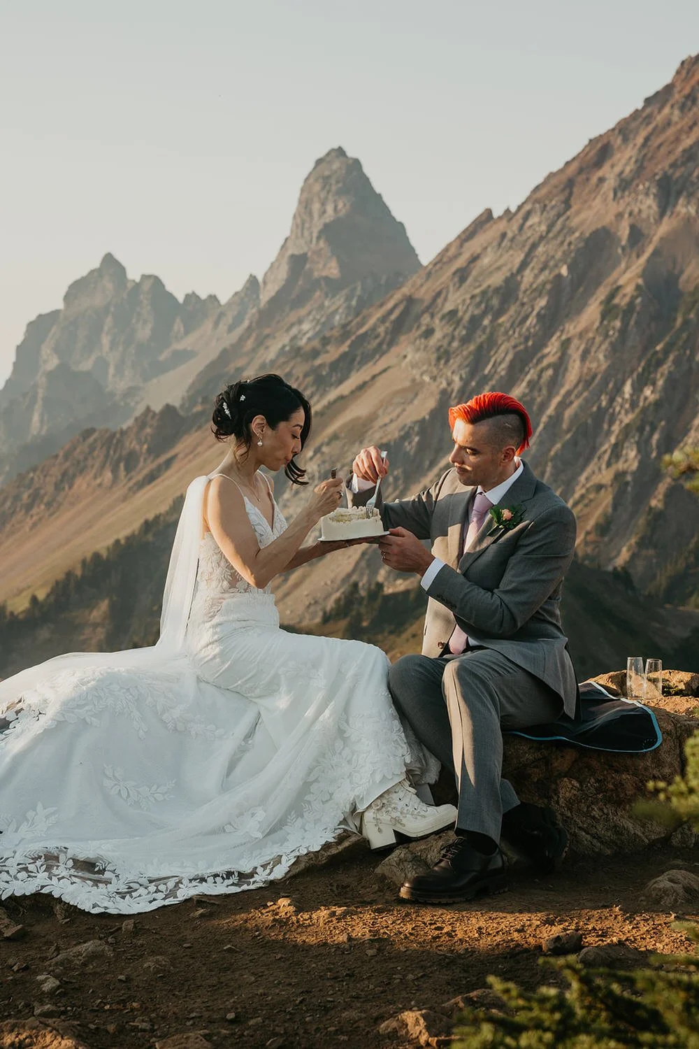 Couple sitting together in the Washington mountains sharing cake during their elopement, with jagged peaks and open alpine scenery behind them