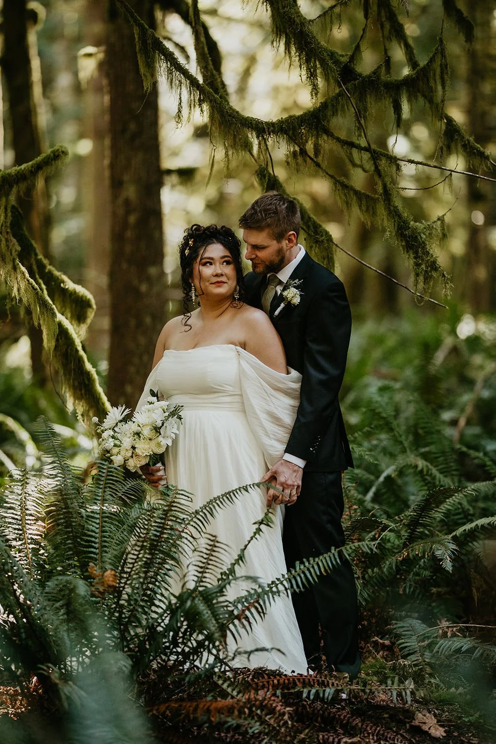 Couple standing close together in a fern-filled forest, moss hanging from branches overhead as light filters through the trees.