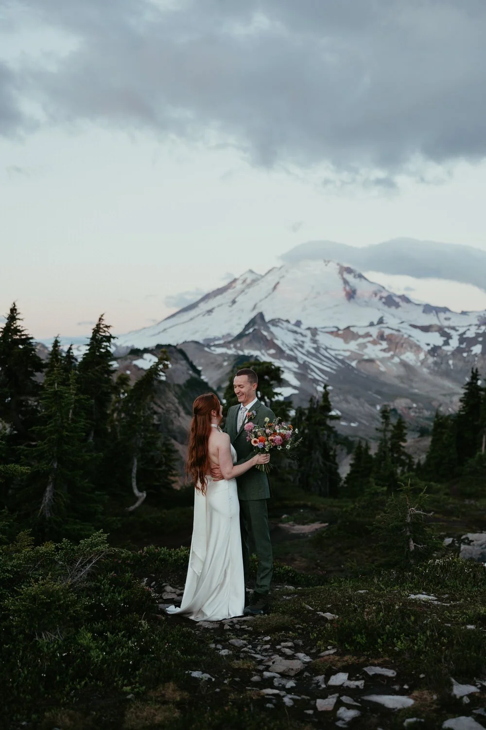 bride and groom stand among rocks and trees with mount baker towering over them in the background