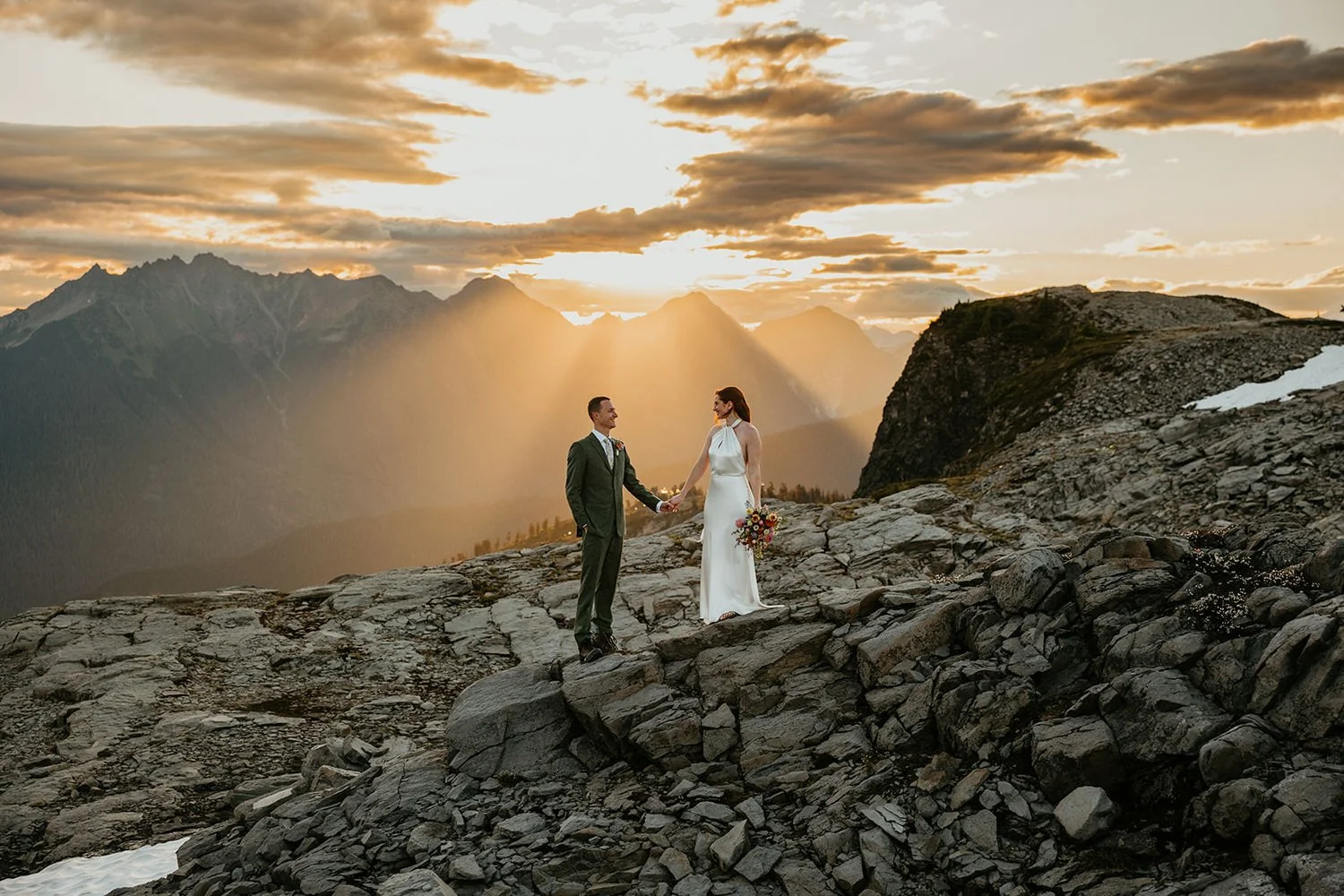 Couple holding hands on a rocky mountain ridge at sunset with dramatic sun rays breaking through clouds over the North Cascades