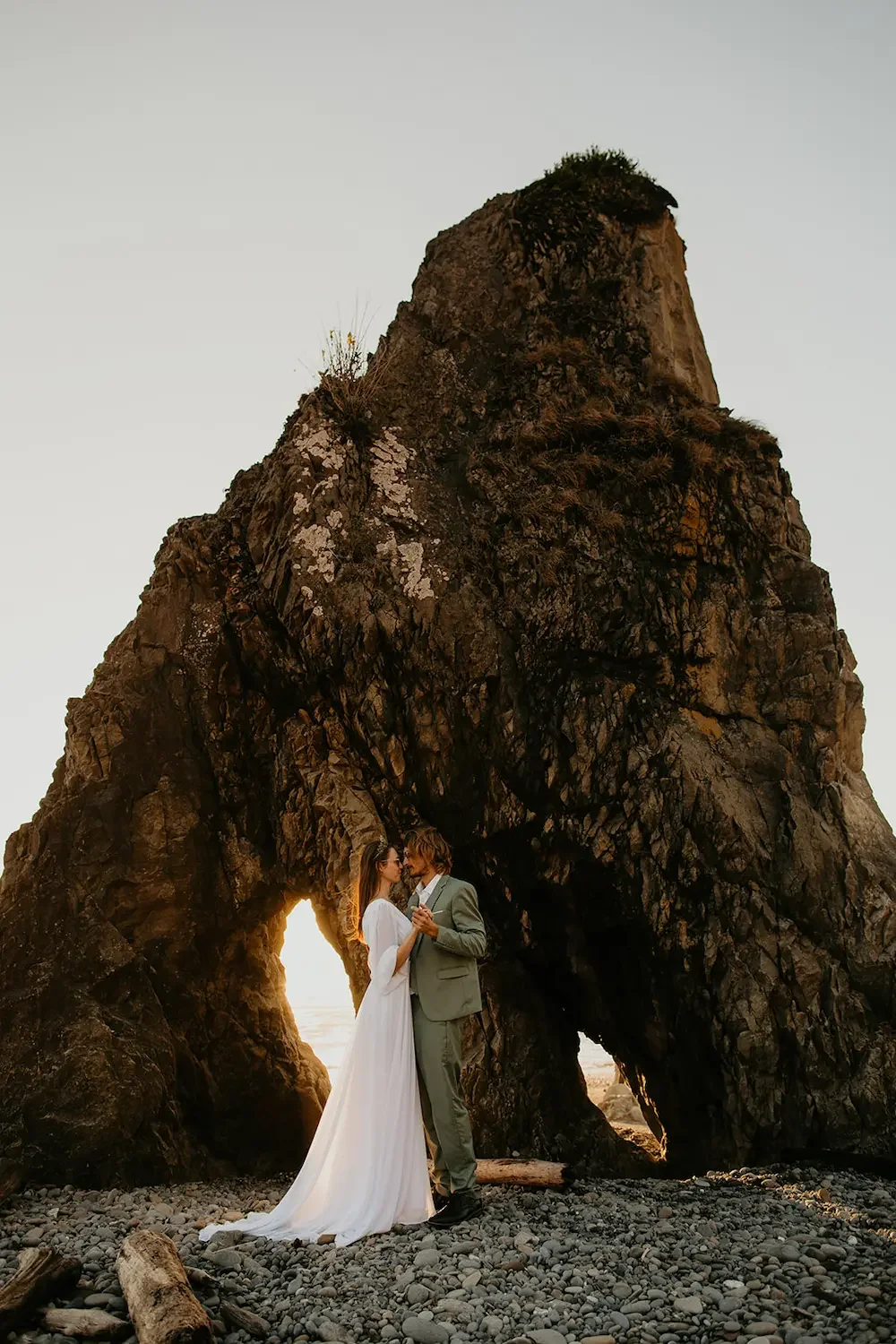 A bride and groom standing close together on a rocky beach at sunset, with a large natural rock arch behind them and the sun setting through the arch.