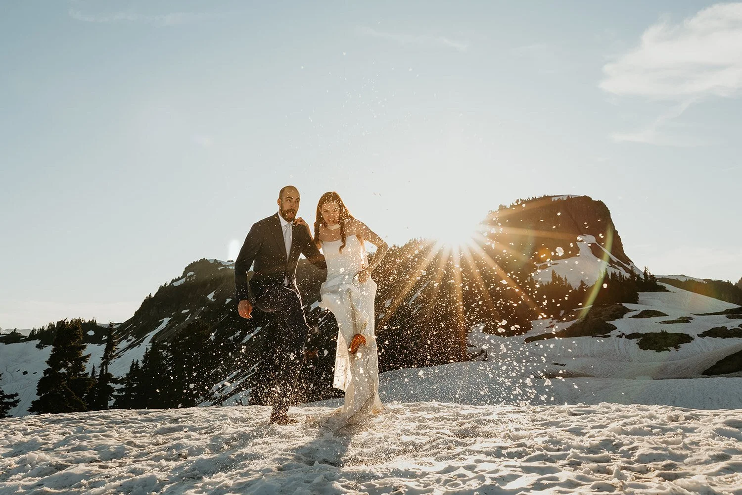Newlyweds running through snow at sunrise in the Washington mountains, bright sunburst peeking over a rocky peak during a winter elopement adventure.