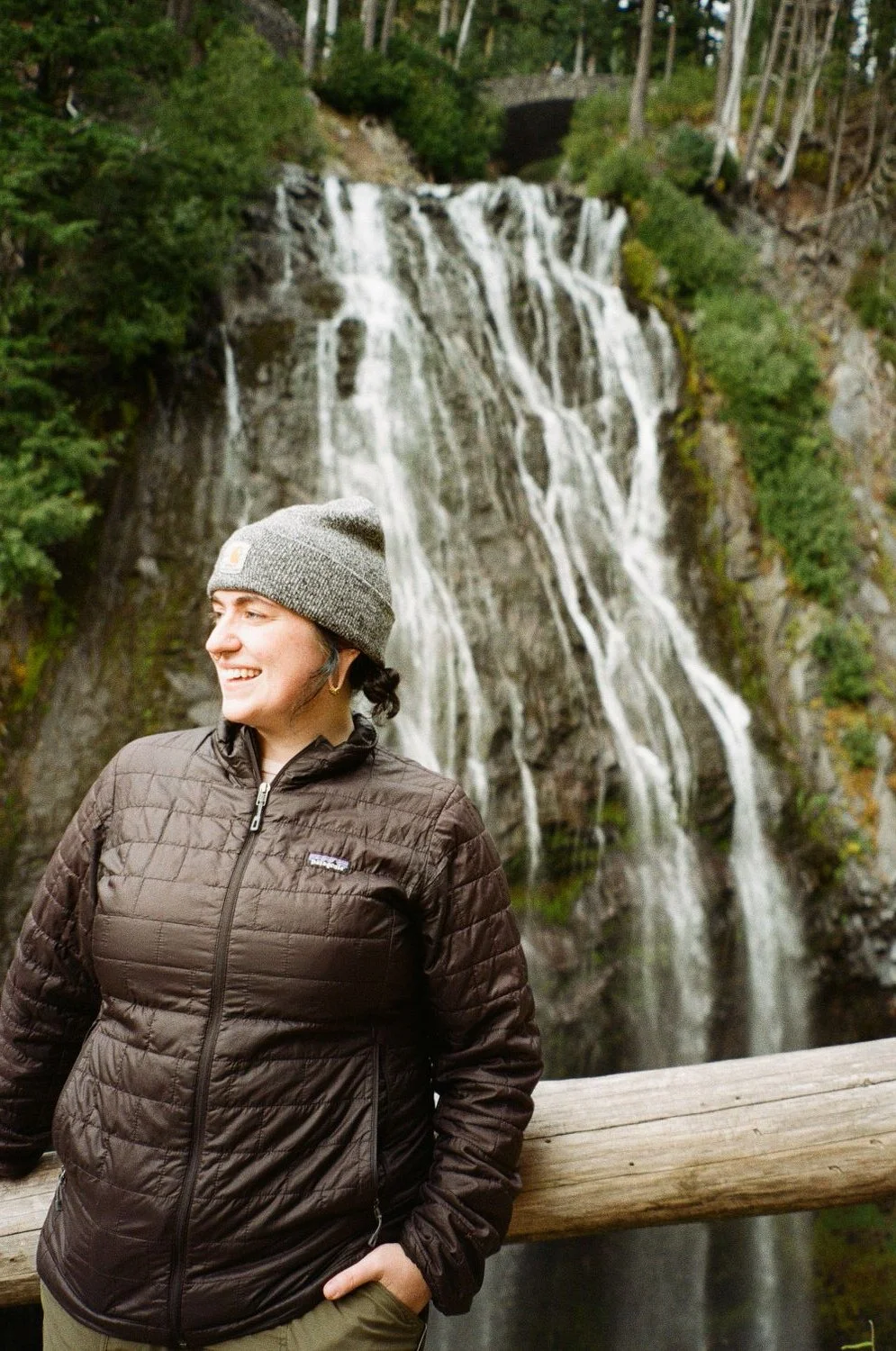 A woman wearing a gray beanie and a brown jacket smiling while standing in front of a waterfall surrounded by trees.