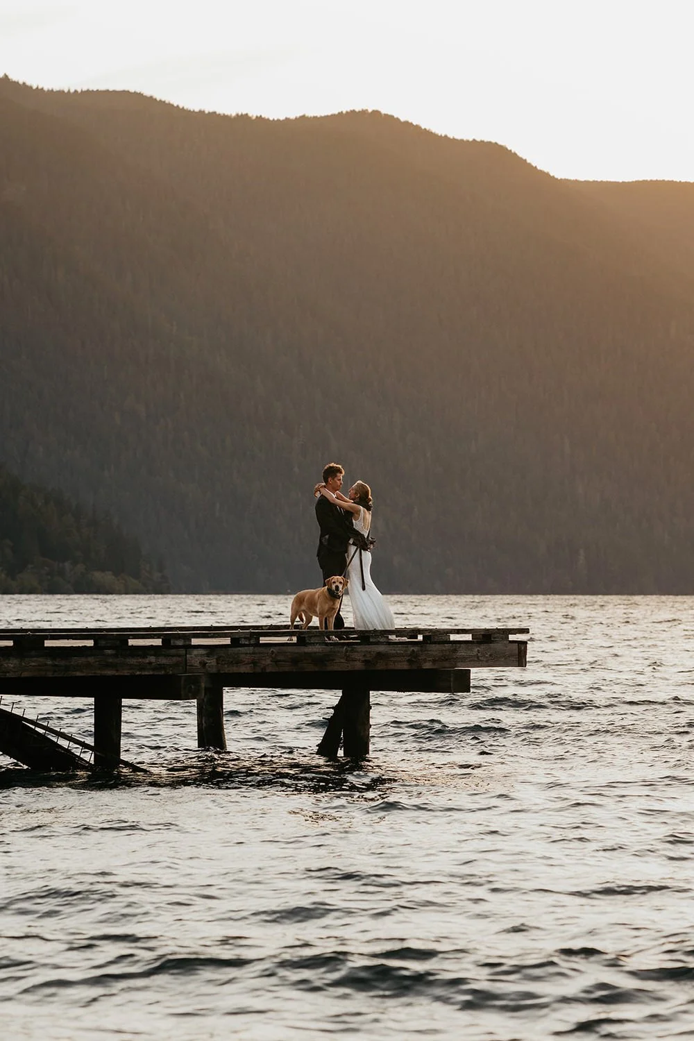 Eloping couple standing on a wooden dock at a mountain lake with their dog, framed by forested hills and calm evening light.