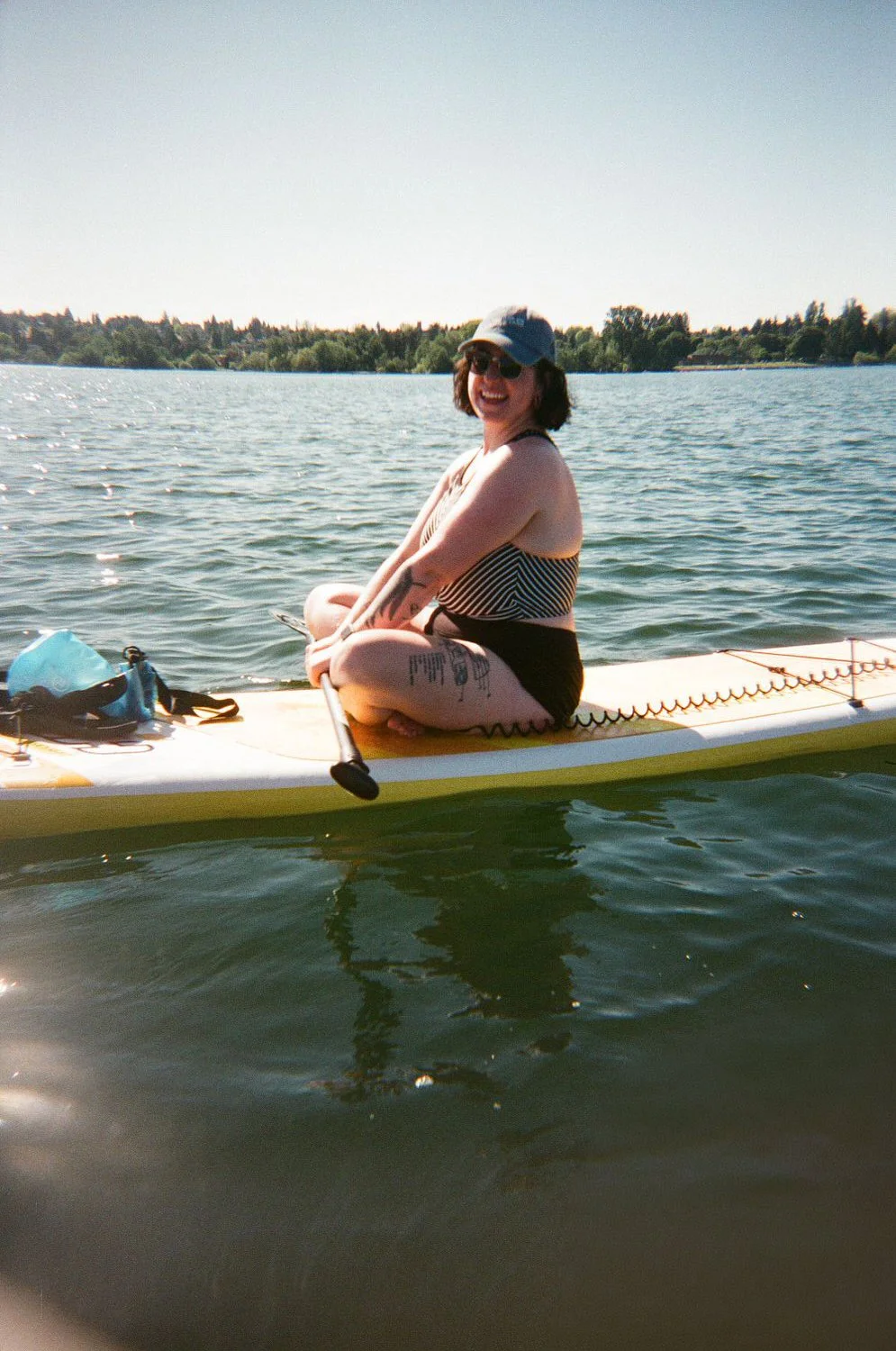 Woman sitting on a paddleboard on a calm lake, smiling, wearing a striped swimsuit, cap, and sunglasses with a background of trees and clear sky.