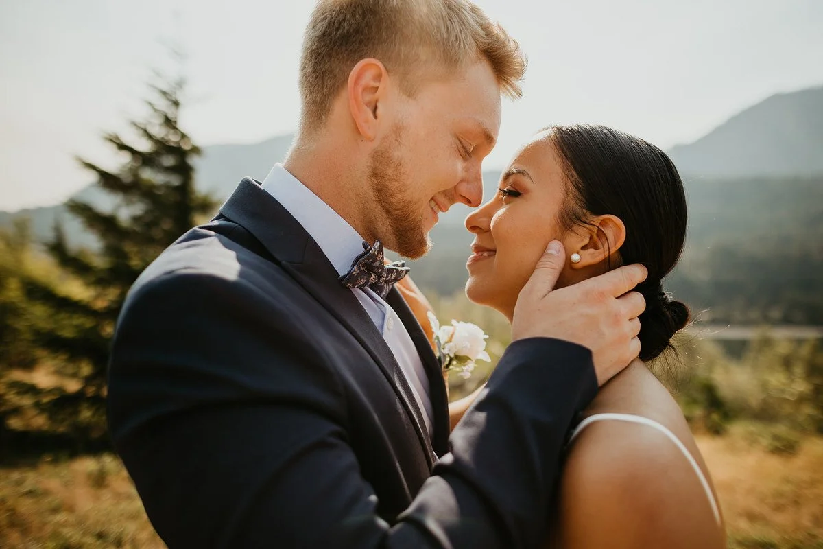 Groom gently holding bride’s face as they smile at each other outdoors with soft evening light and distant mountain scenery.
