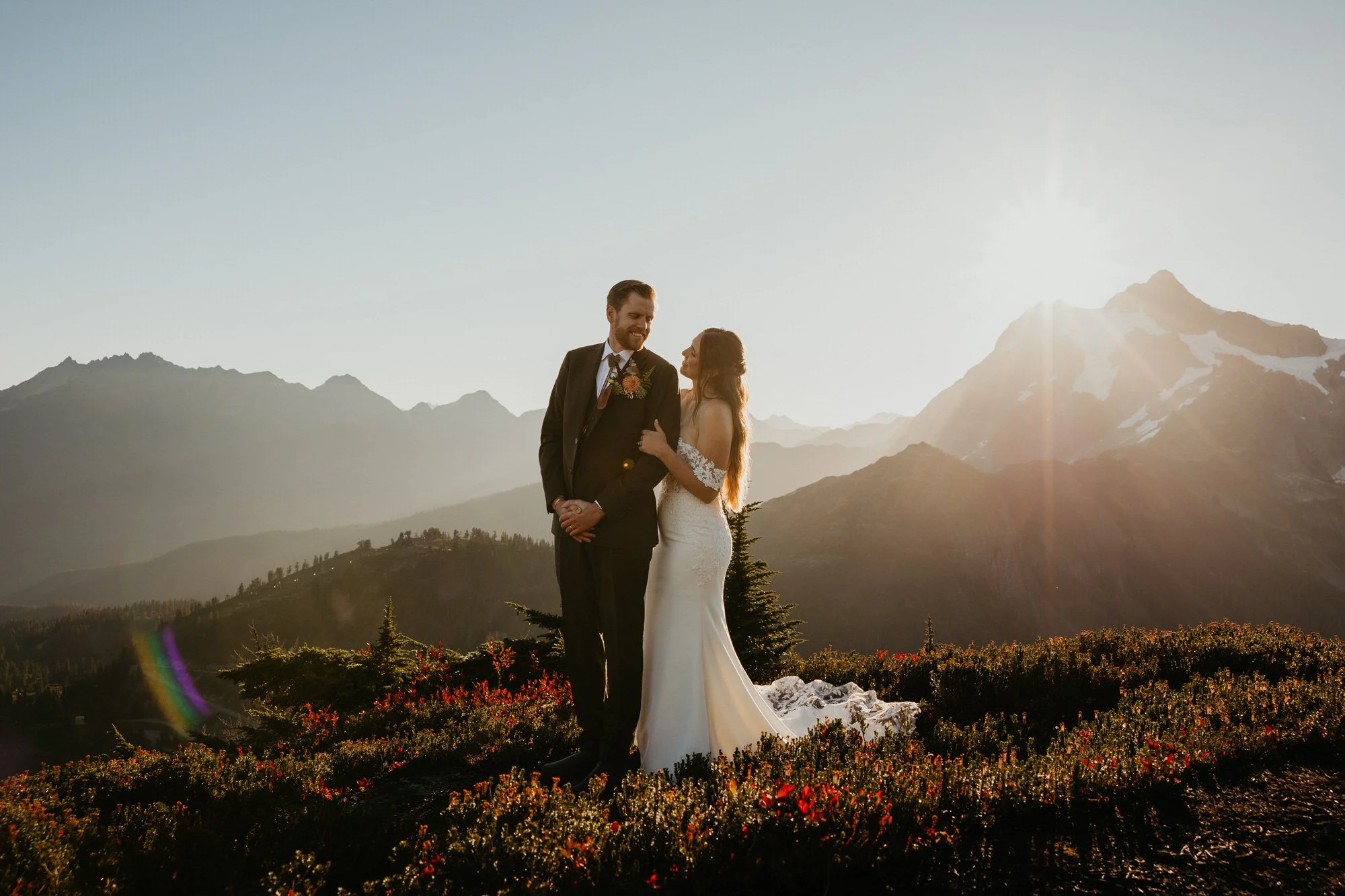 A bride and groom stand together outdoors during sunset, with mountains in the background and vibrant plants in the foreground.