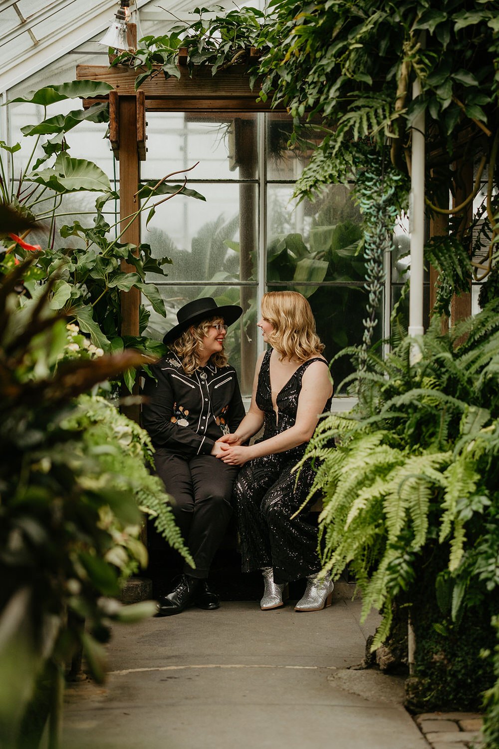 Two brides holding hands and smiling at each other inside a lush greenhouse, surrounded by greenery and natural light.