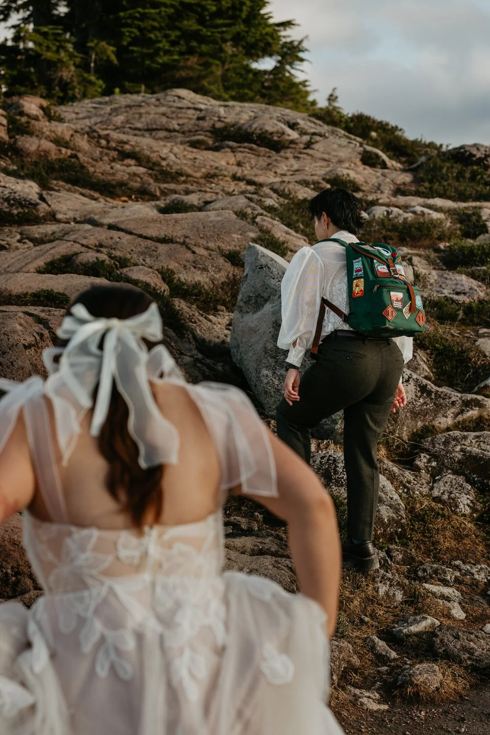 Two brides climbing over a rocky slope together, one wearing a backpack covered in trail patches