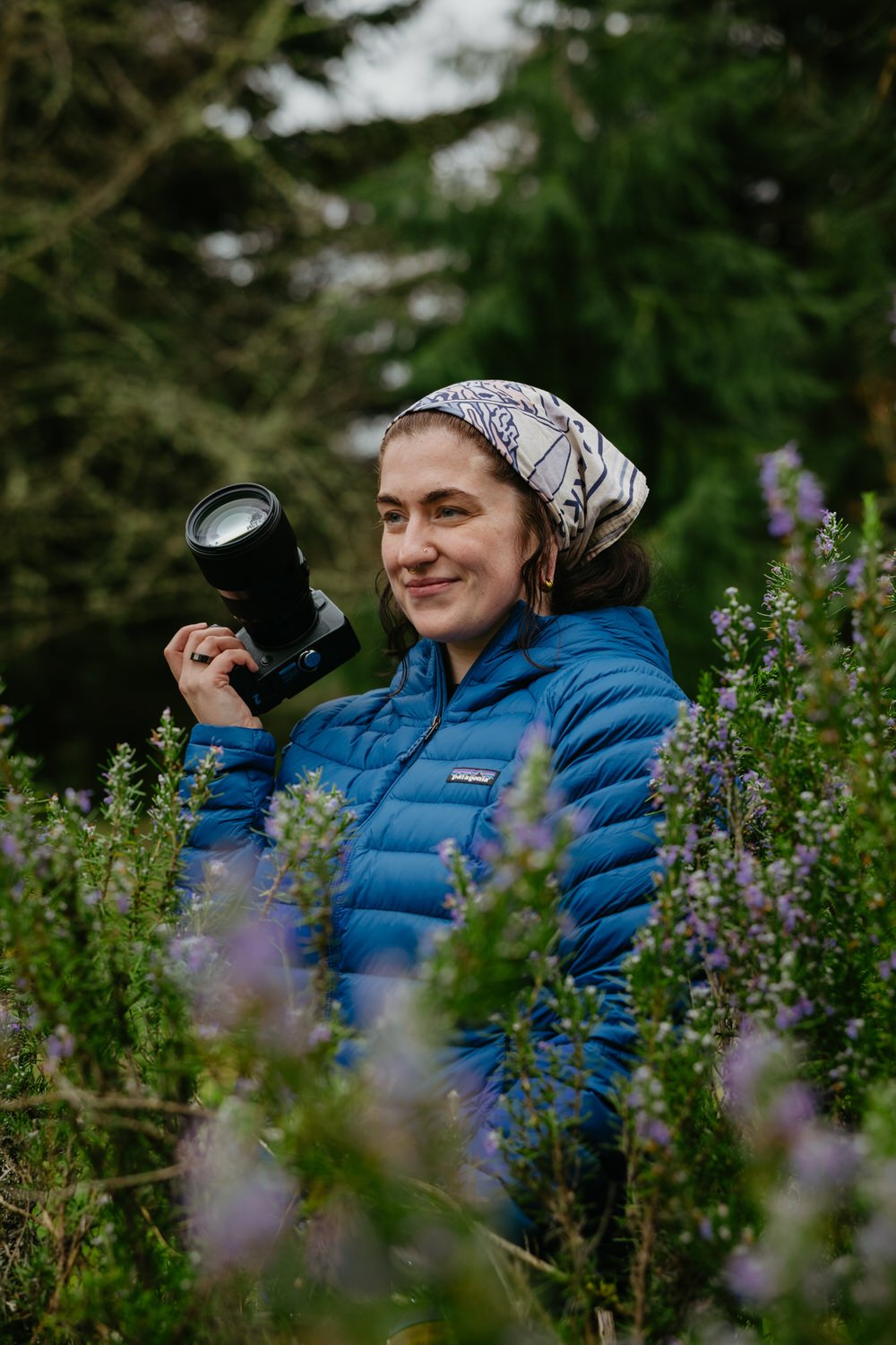Van Gachnang stands among a purple rosemary bush wearing a blue jacket and holding a camera