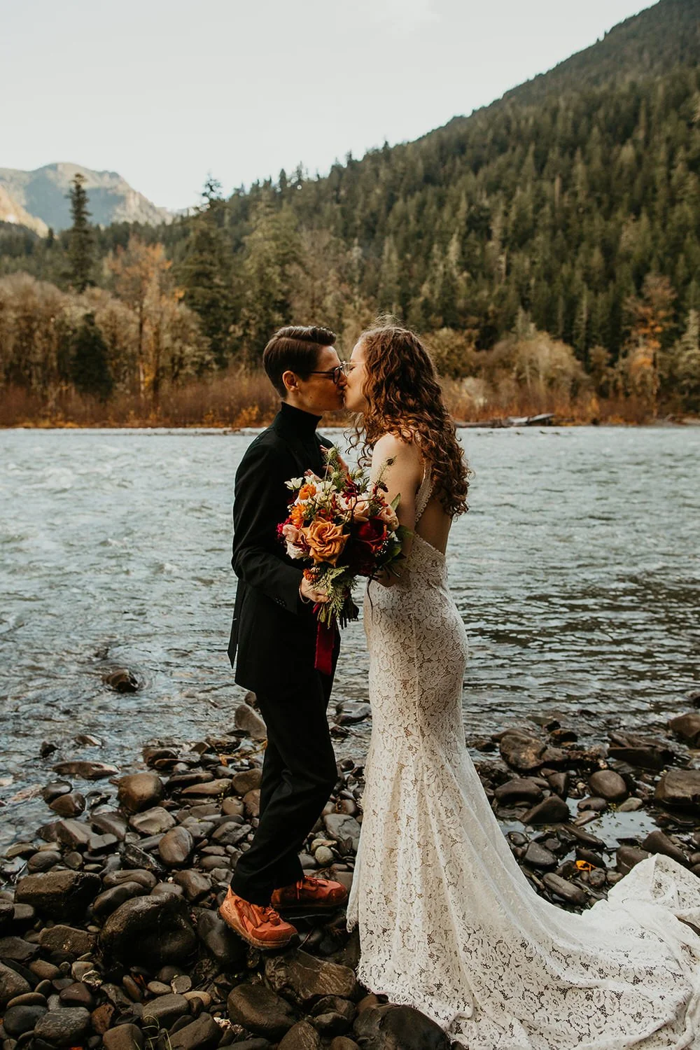Two brides sharing a kiss on a rocky riverbank with forested mountains rising behind them during an intimate outdoor elopement.