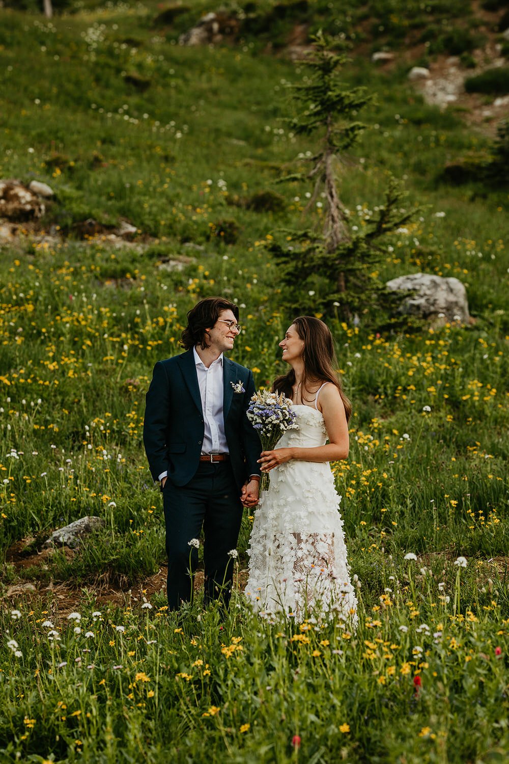 A couple in wedding attire walks hand in hand through a wildflower-filled meadow surrounded by lush green hillsides.