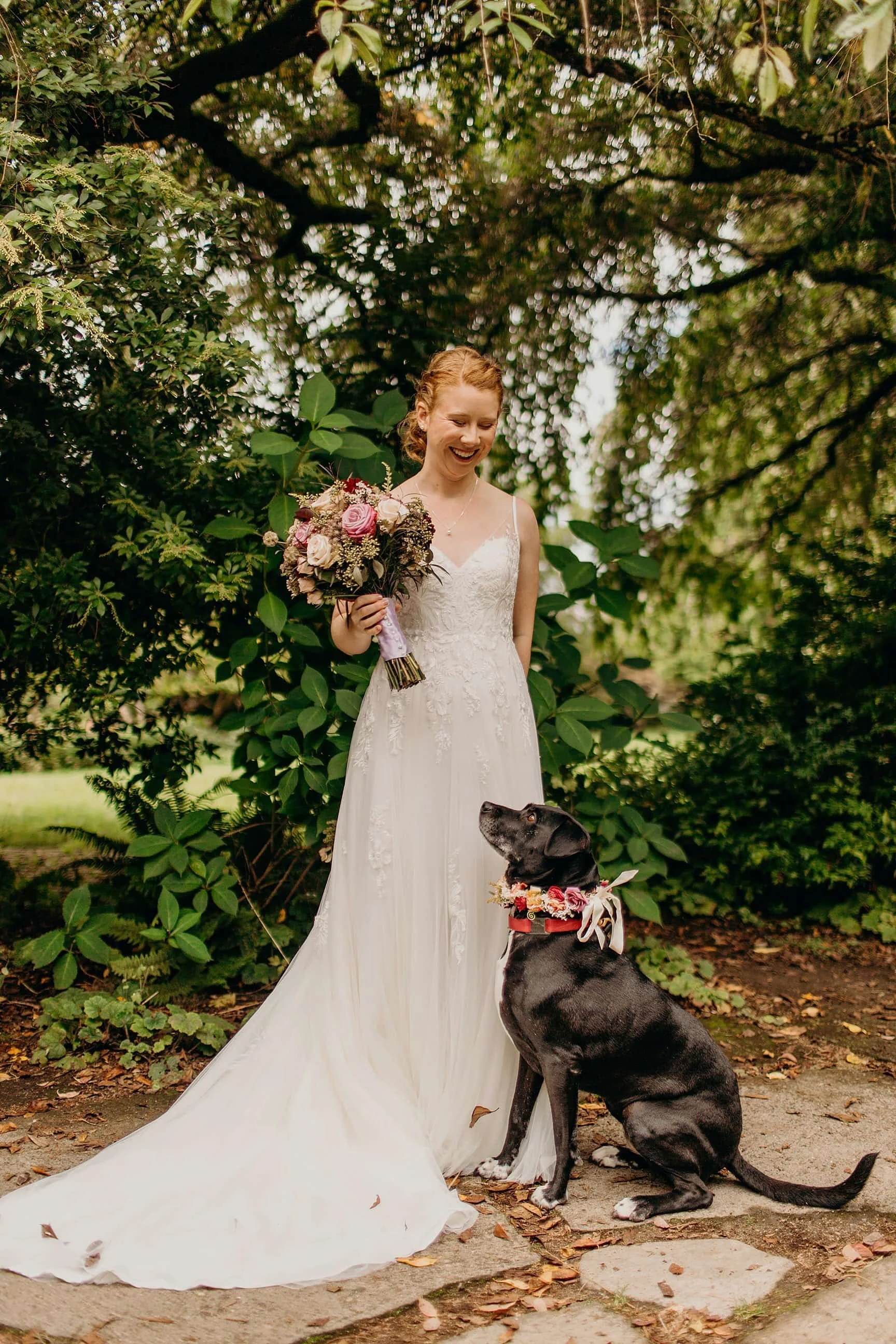 Bride holding a bouquet and smiling at her black dog wearing a floral collar in a garden setting