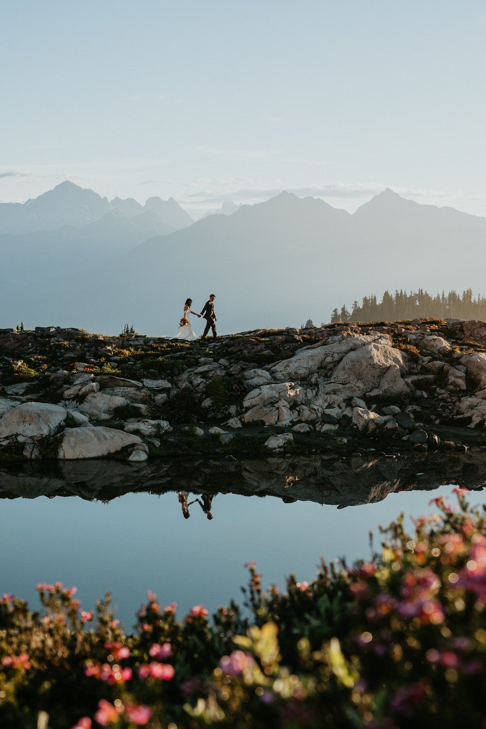 Couple walking along a rocky lakeside ridge with pink wildflowers blooming in the foreground and distant peaks beyond