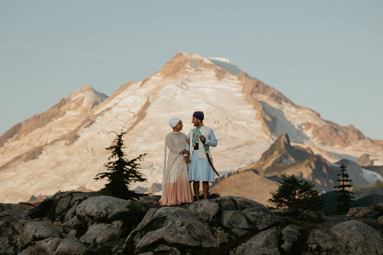 Couple in traditional Sikh wedding attire standing on a rocky overlook with Mount Baker rising behind them during golden hour in Washington.