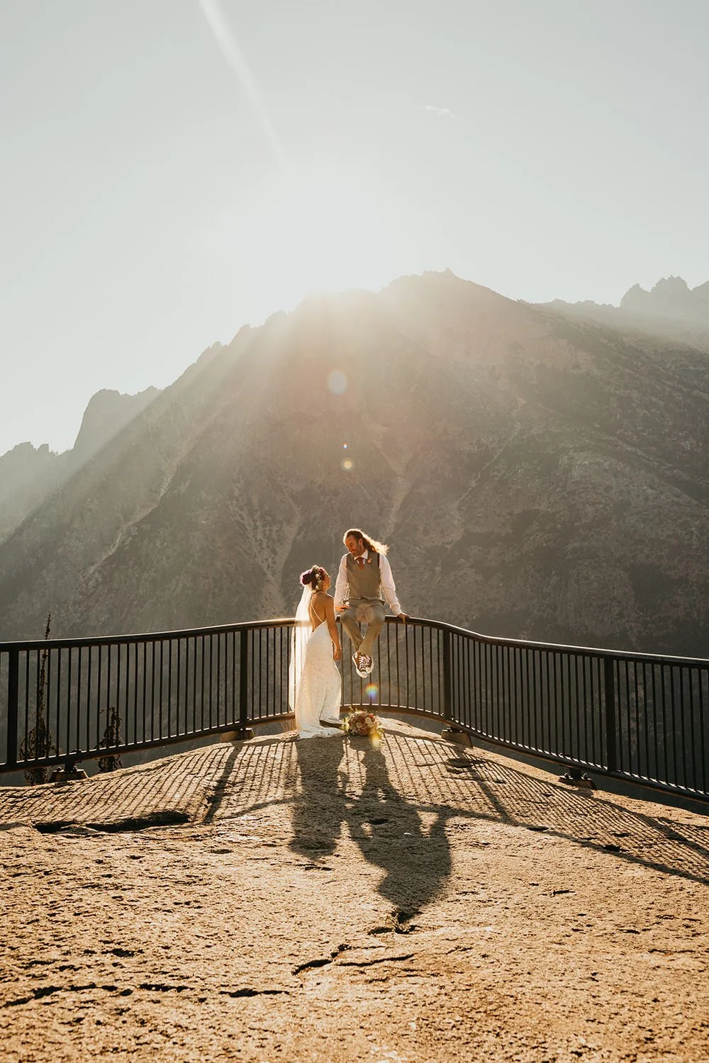 Groom sits on railing edge in front of North Cascades peaks while bride looks up at him from the ground
