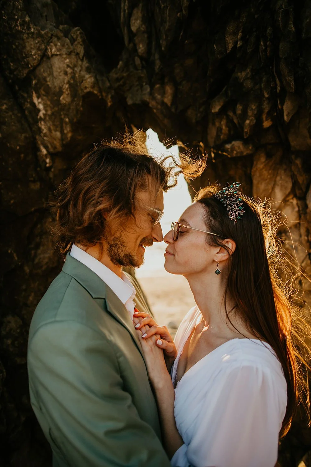 Close-up of a couple standing beneath a rocky sea arch at golden hour, sunlight glowing through the opening behind them.