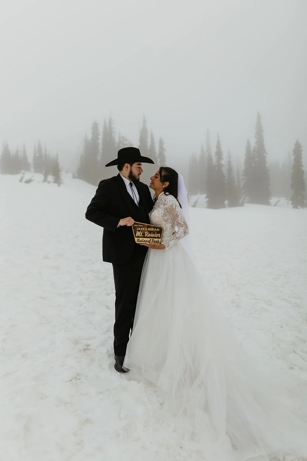 Snowy mountain elopement with couple holding a Mount Rainier National Park sign in foggy winter conditions